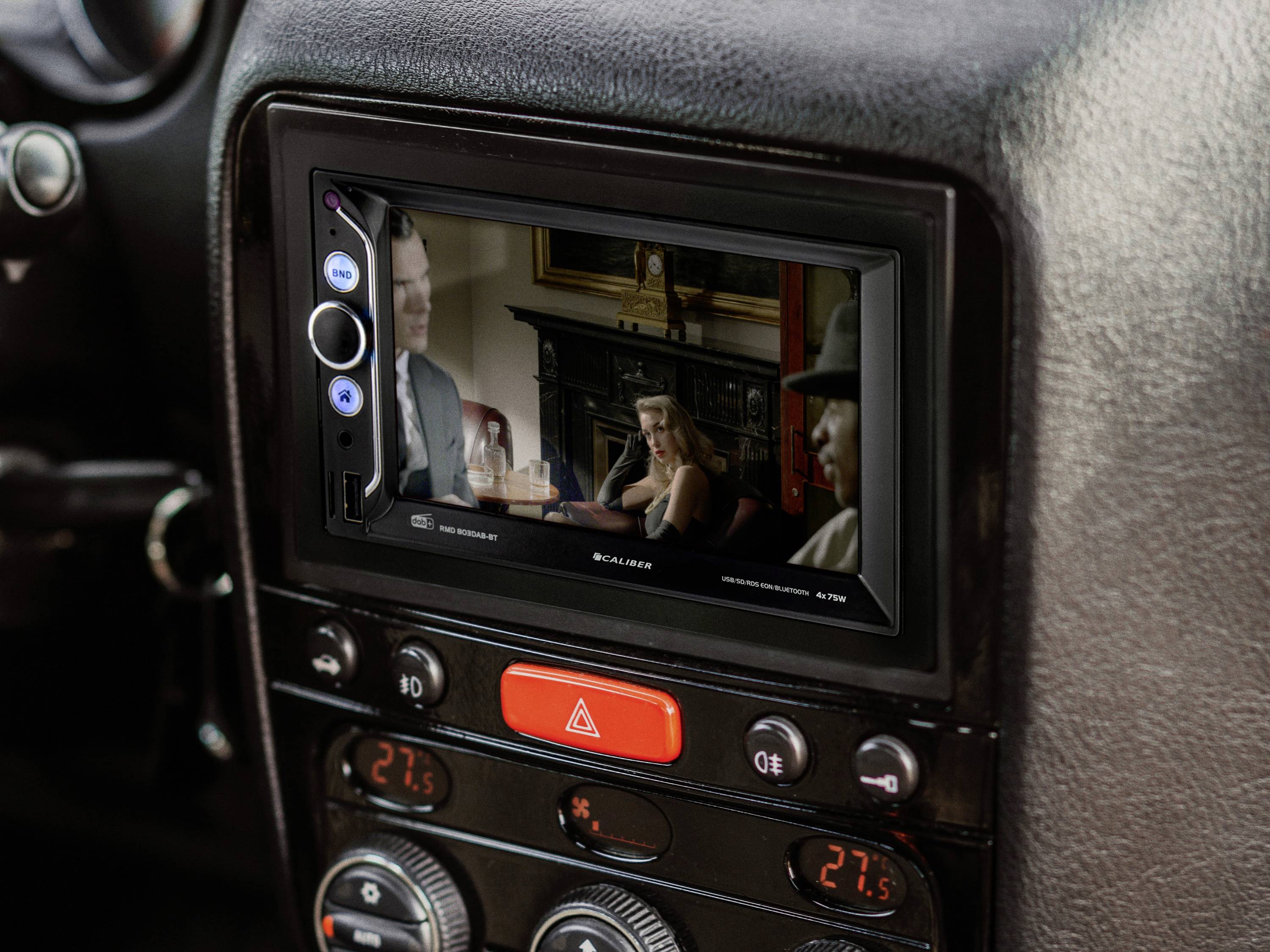 Bildschirm eines Auto-Entertainment-Systems zeigt eine Filmszene mit drei Personen in einem Raum. Es sind Heizungskontrollen sichtbar.