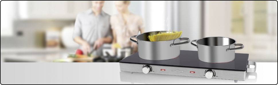 Zwei Kochtöpfe stehen auf einem Elektrokochfeld, im Hintergrund unscharf zwei Personen beim Kochen in einer modernen Küche.