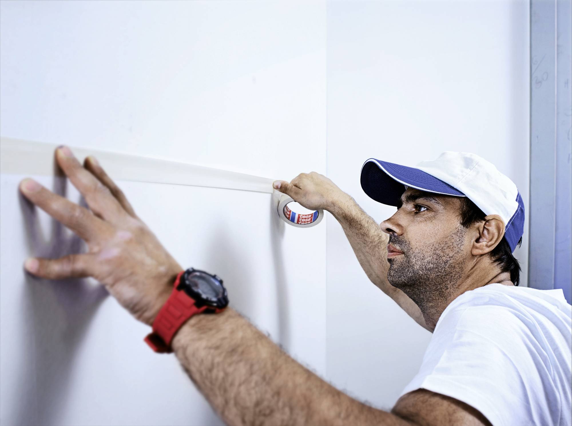 Ein Mann klebt ein Maßband an eine Wand, trägt ein weißes T-Shirt und eine Kappe. Er konzentriert sich auf präzises Arbeiten.