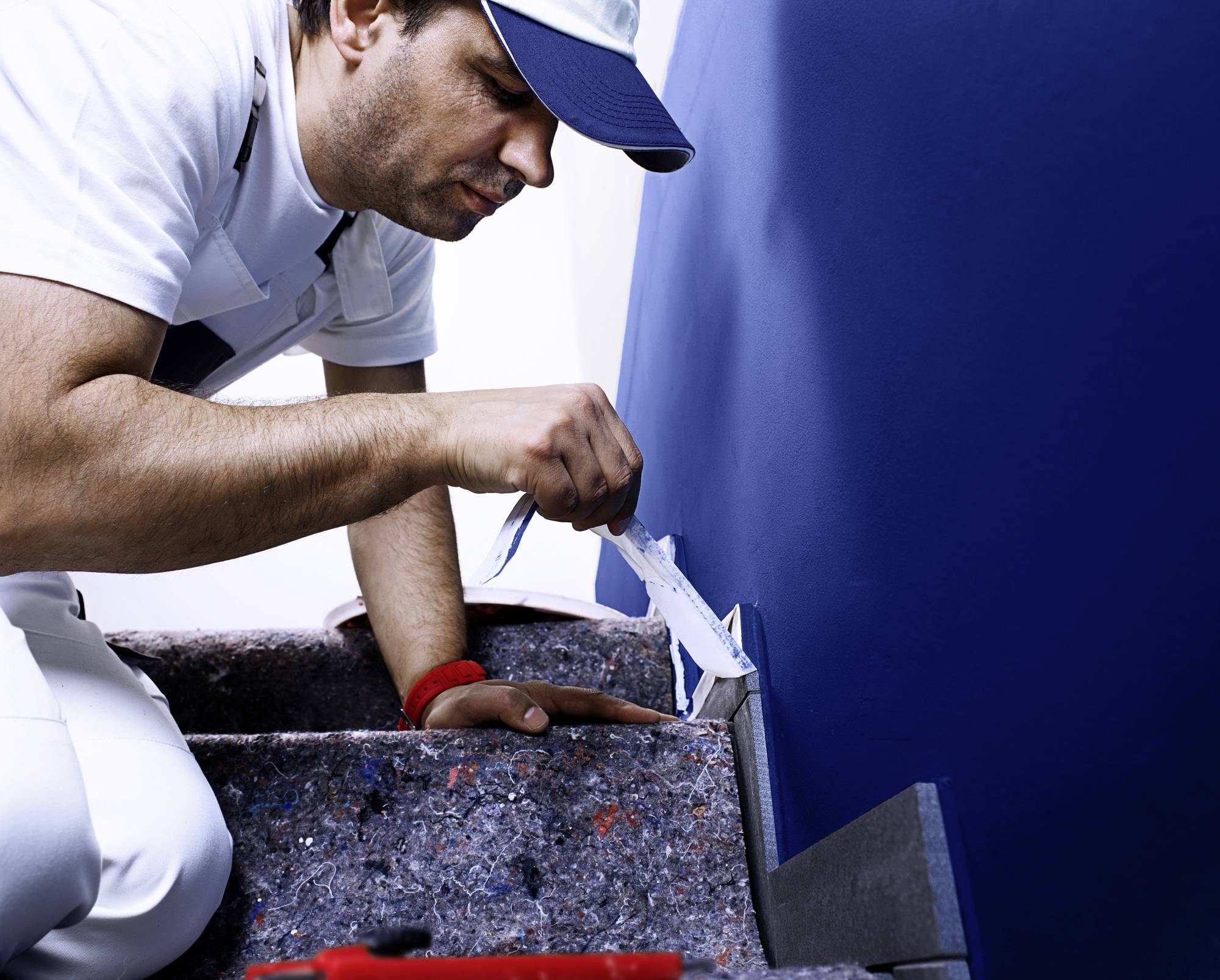 Ein Handwerker streicht eine Wand blau mit einem Pinsel. Er trägt weiße Arbeitskleidung und eine blaue Kappe.
