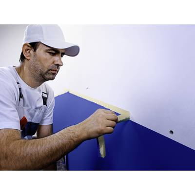 Ein Maler in Arbeitskleidung klebt mit einem Klebeband eine Wand ab, um diese zweifarbig zu streichen. Die Wand ist teilweise blau.