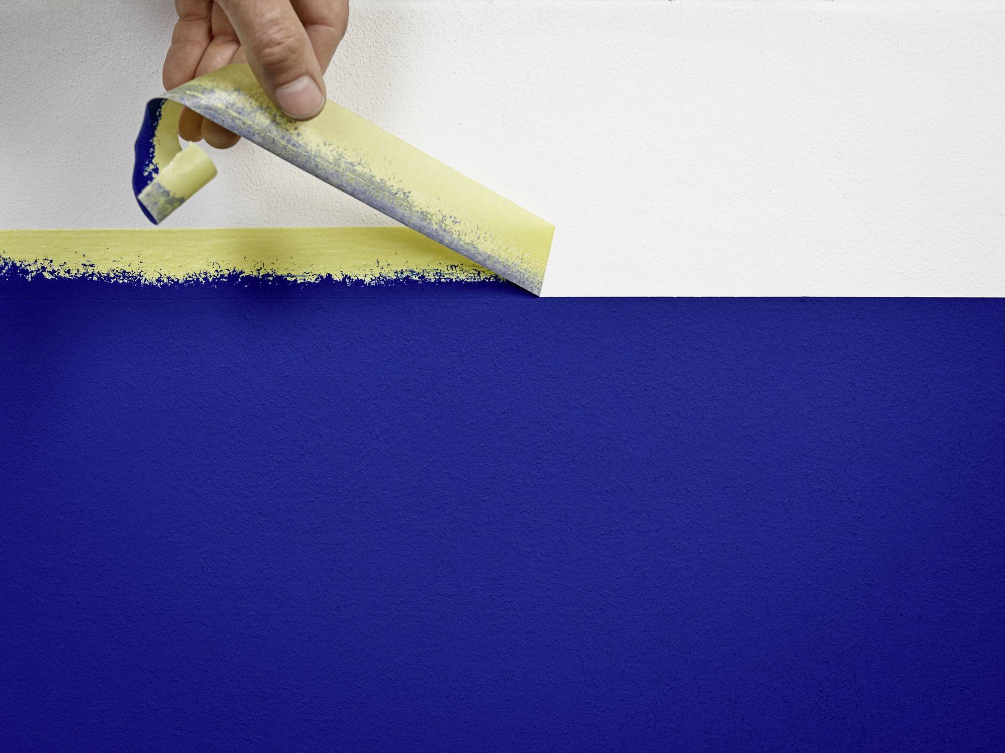 Eine Hand zieht Klebeband von einer blau-weiß gestrichenen Wand ab, wodurch eine gerade Kante zwischen den Farben entsteht.
