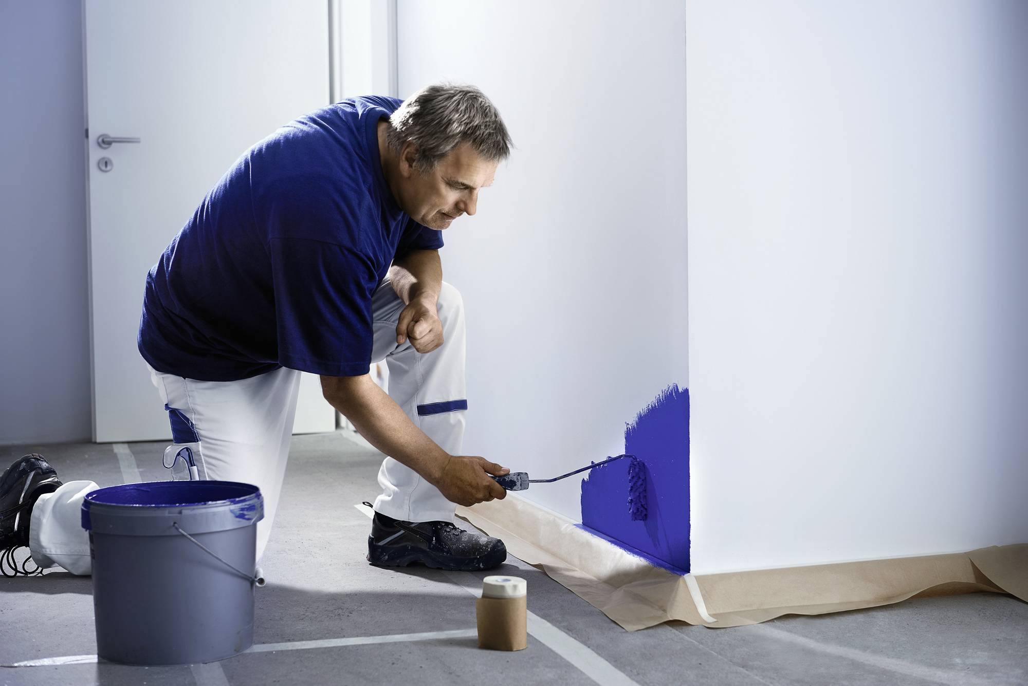 Ein Mann kniet und streicht eine Wand mit blauer Farbe. Der Boden ist mit Abdeckpapier geschützt. Ein Eimer Farbe steht neben ihm.