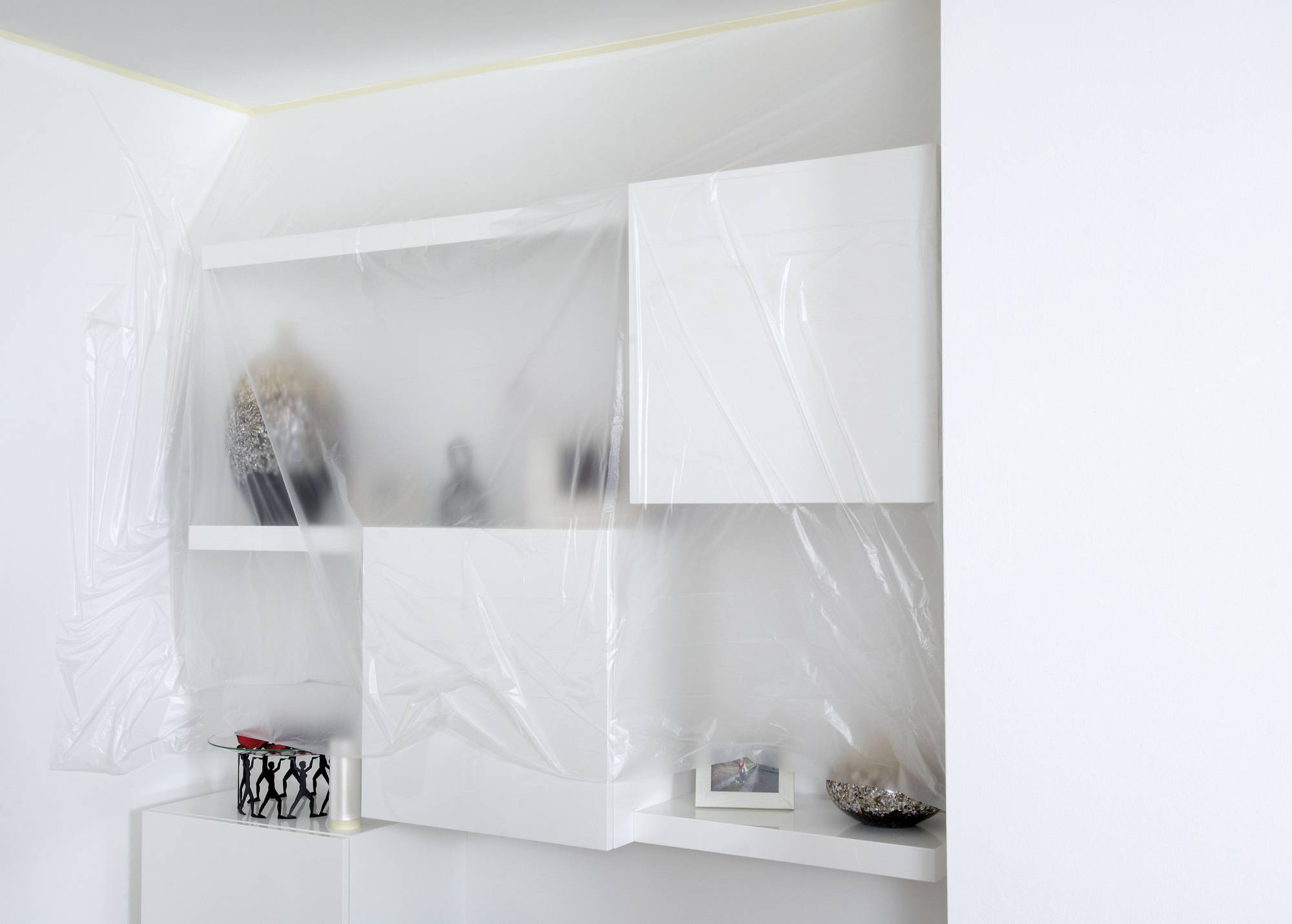 Ein weißes Regal mit Vasen und einem eingerahmten Foto, abgedeckt mit transparenter Plastikfolie in einem modern gestalteten Raum.