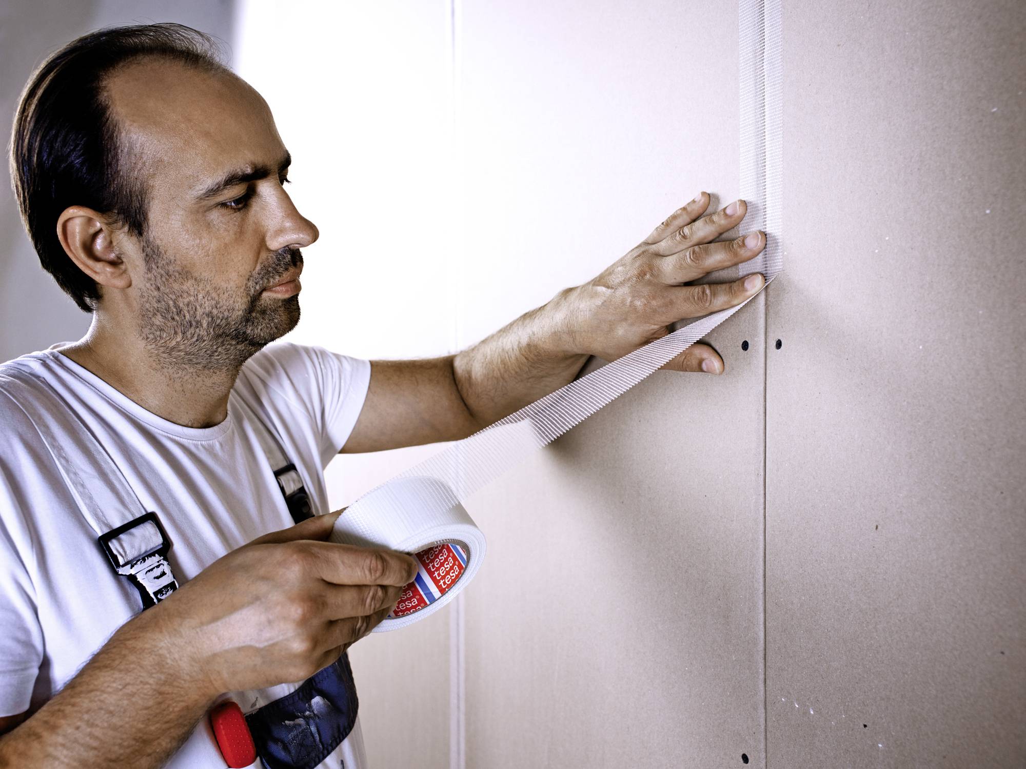 Ein Mann befestigt Trockenbauplatten an einer Wand mit Klebeband, trägt Malerbekleidung und konzentriert sich auf seine Arbeit.