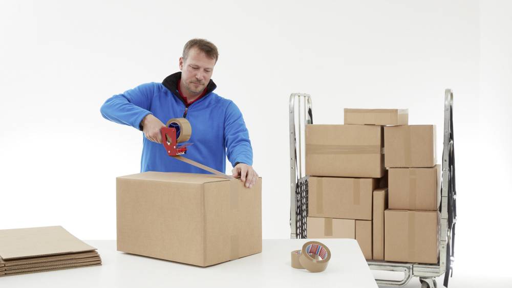 Ein Mann in blauem Pullover verpackt einen Karton mit Klebeband. Daneben stehen mehrere gestapelte Kartons auf einem Wagen.