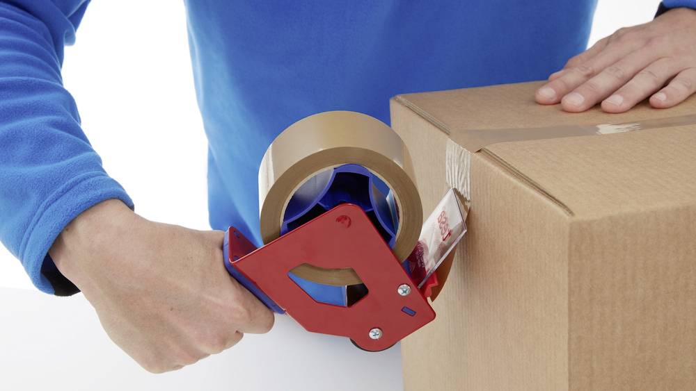 Person verpackt einen Karton mit Klebeband. Eine Hand hält einen roten Abroller, die andere stützt die Schachtel.