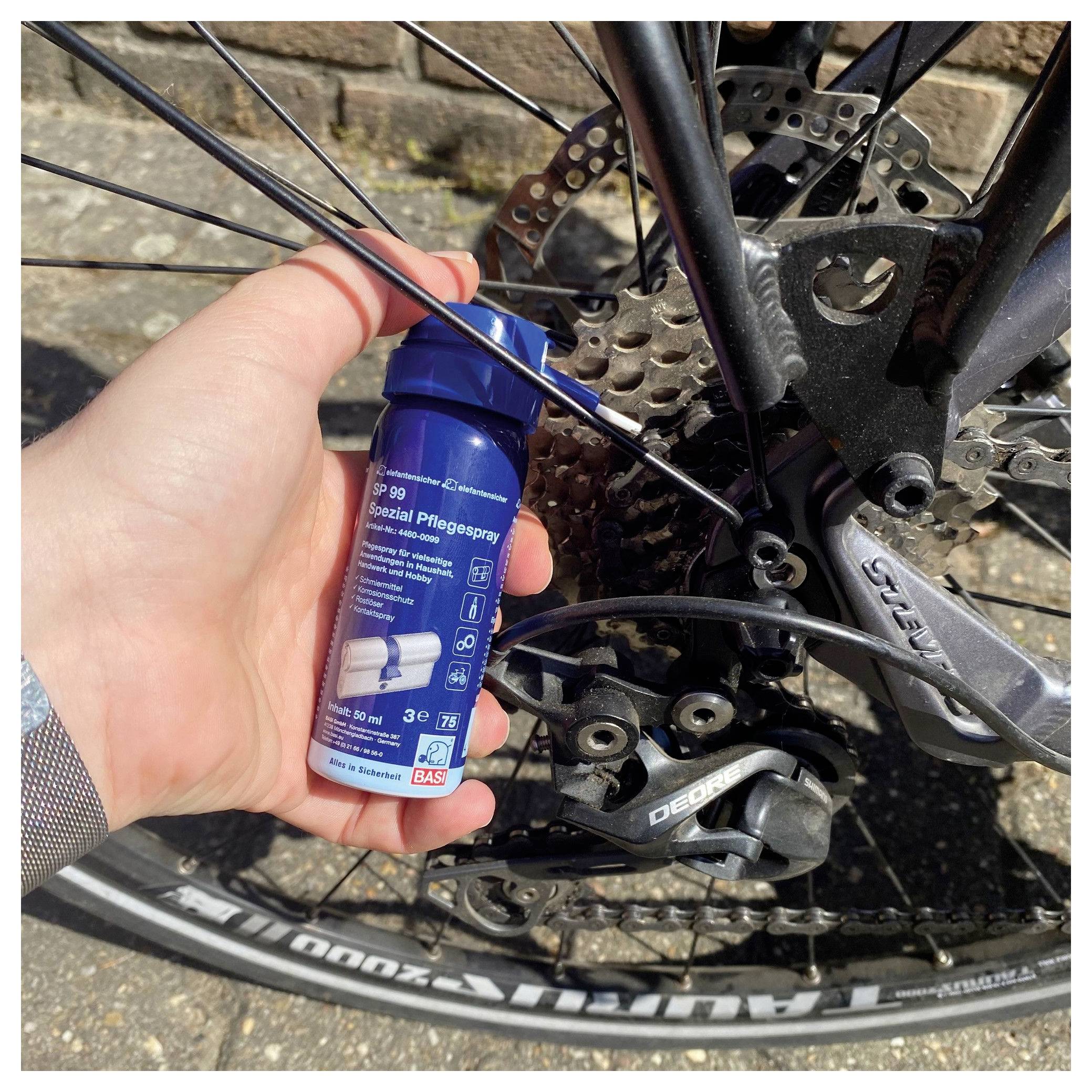 Eine Hand hält eine blaue Sprühdose in der Nähe einer Fahrradkette und eines Schaltwerks. Die Dose ist mit Reinigungsmittelinformationen in verschiedenen Sprachen gekennzeichnet.