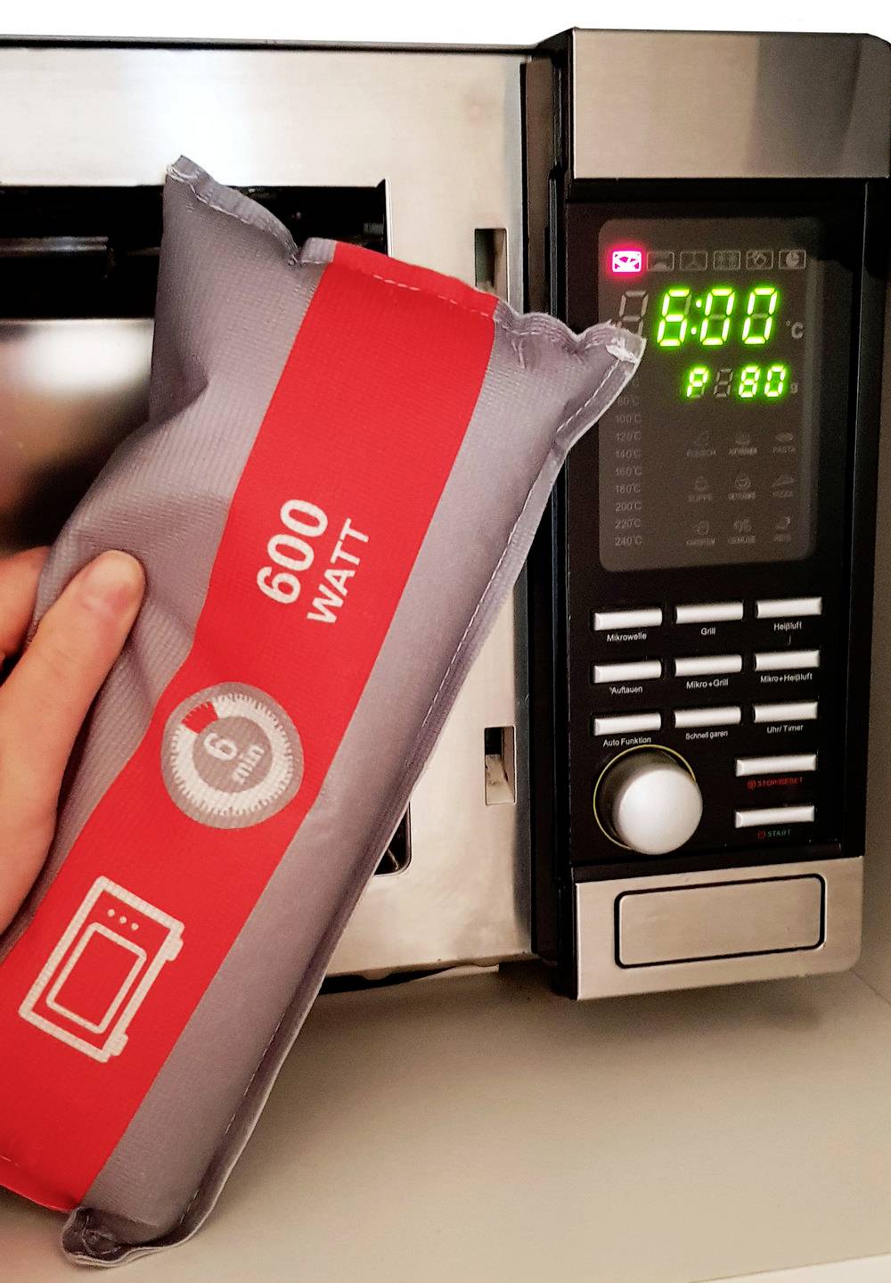Eine Hand hält eine Wärmekissen-Verpackung vor eine Mikrowelle. Die Anzeige zeigt 6 Minuten und 800 Watt.