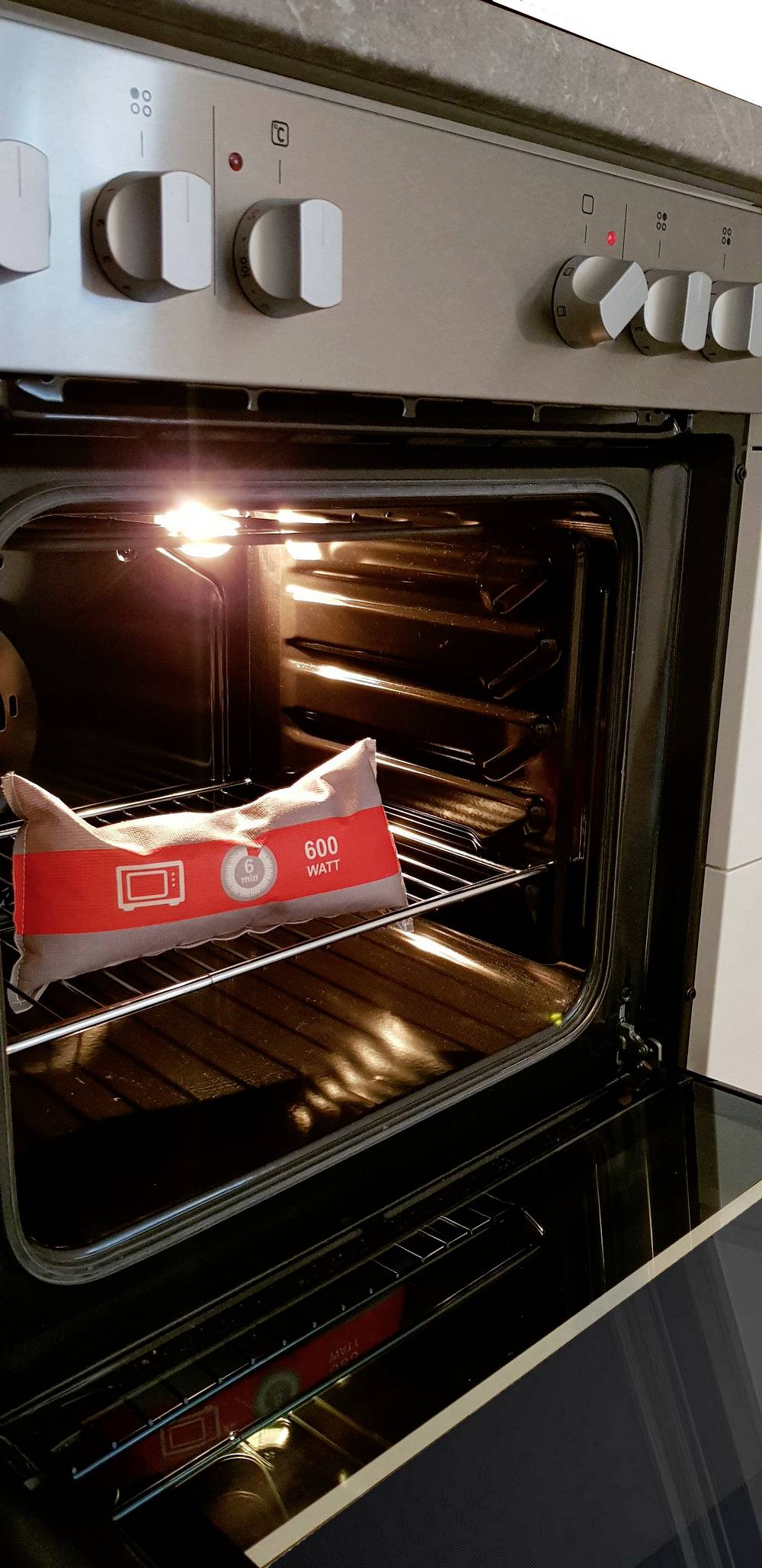 Ein geöffneter Backofen mit eingeschaltetem Licht. Auf dem Rost liegt ein rotes Kissen mit Symbolen und der Zahl '600 Watt'.