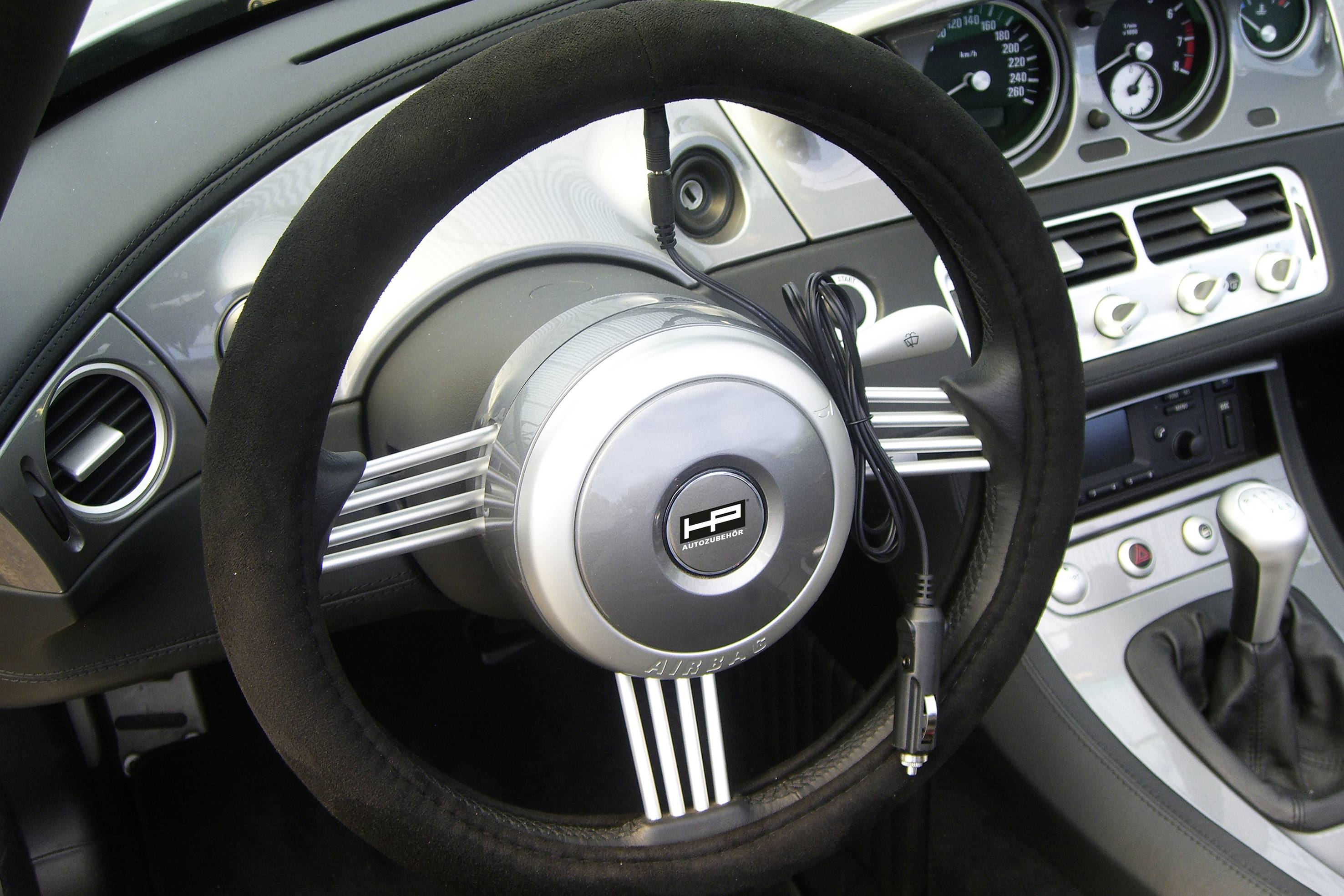 Lenkrad eines Autos mit mittig platziertem Logo auf einer metallischen Oberfläche. Schaltknauf und Armaturenbrett im Hintergrund ersichtlich.