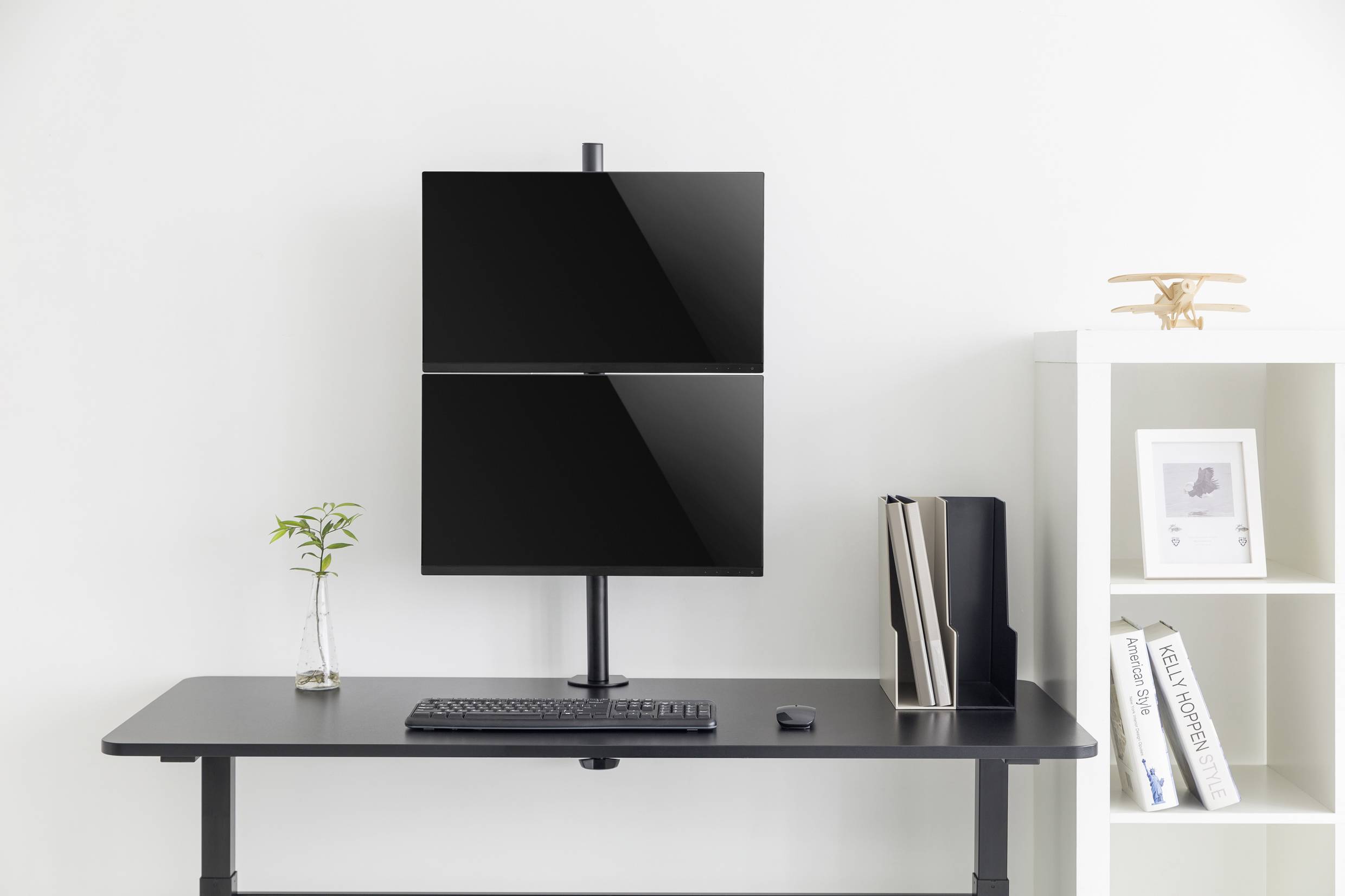 Ein minimalistischer Schreibtisch mit zwei gestapelten Monitoren, einer Tastatur, einem Mausgerät, einer Vase mit Pflanze und Bücherregalen.