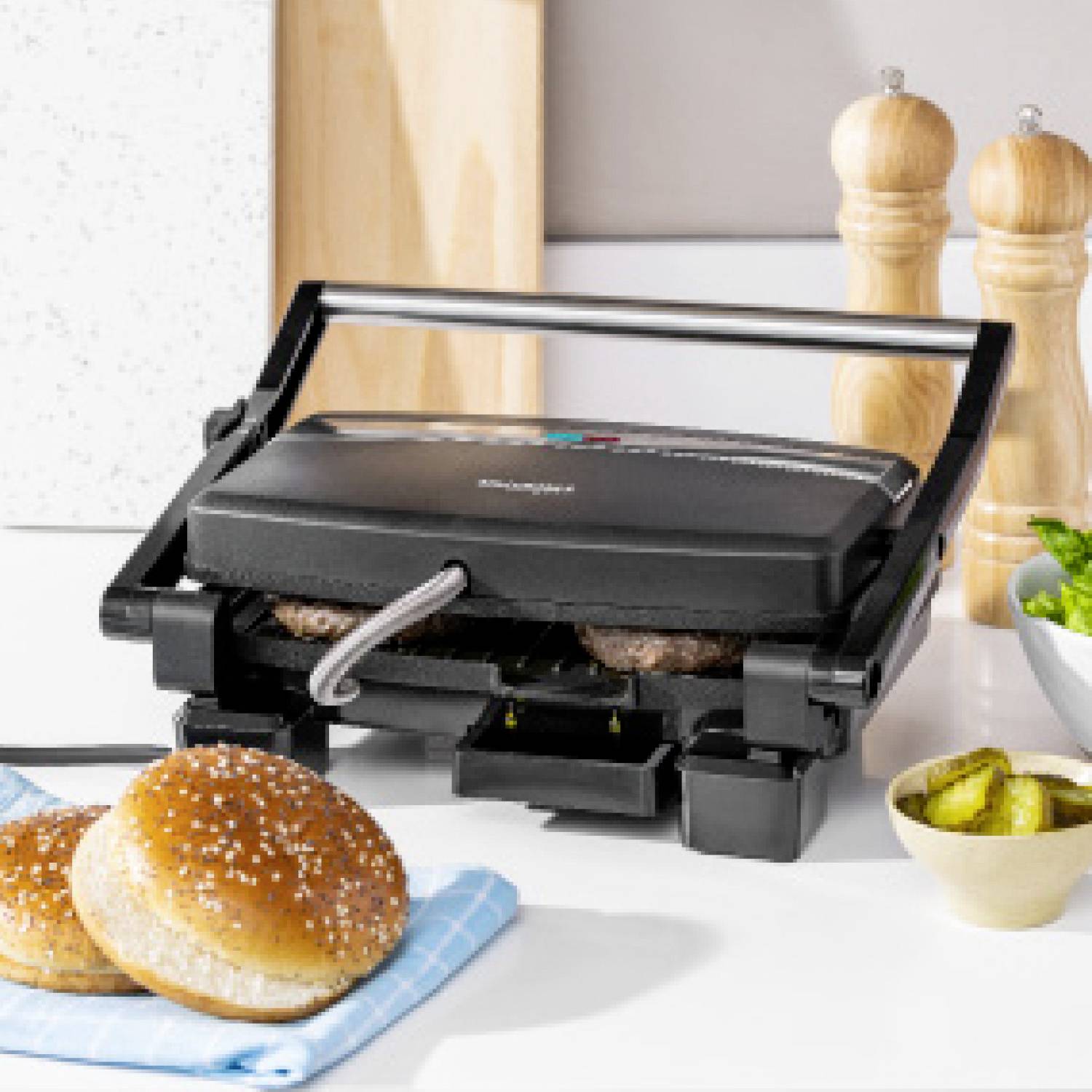 Ein Kontaktgrill auf einer Küchenarbeitsplatte grillt Burger-Patties. Im Hintergrund sind Salz- und Pfefferstreuer sowie eine Schüssel mit Gewürzgurken.