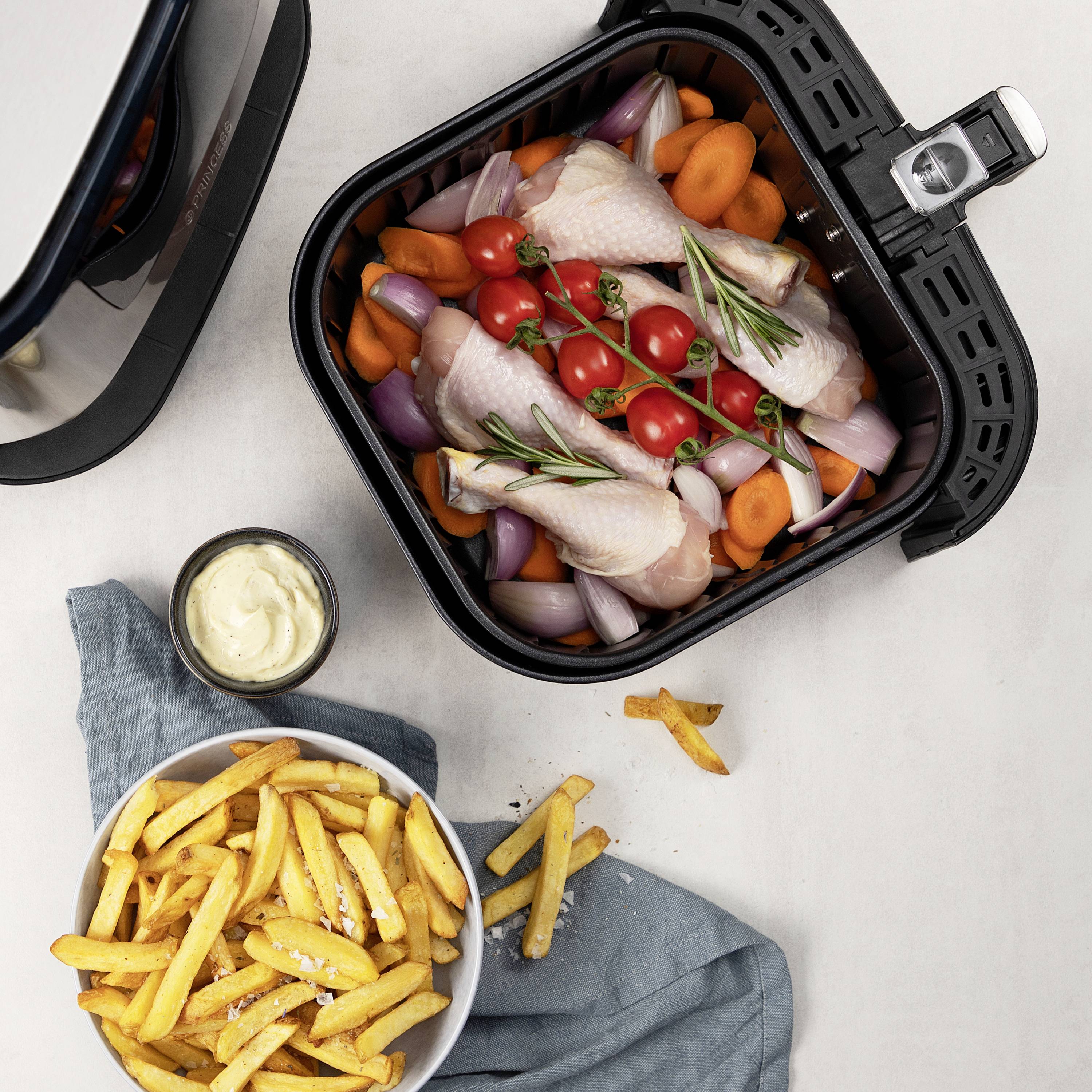 Eine am Tisch stehende Heißluftfritteuse mit Hähnchen, Tomaten und Gemüse im Korb. Daneben stehen Pommes frites in einer Schüssel.