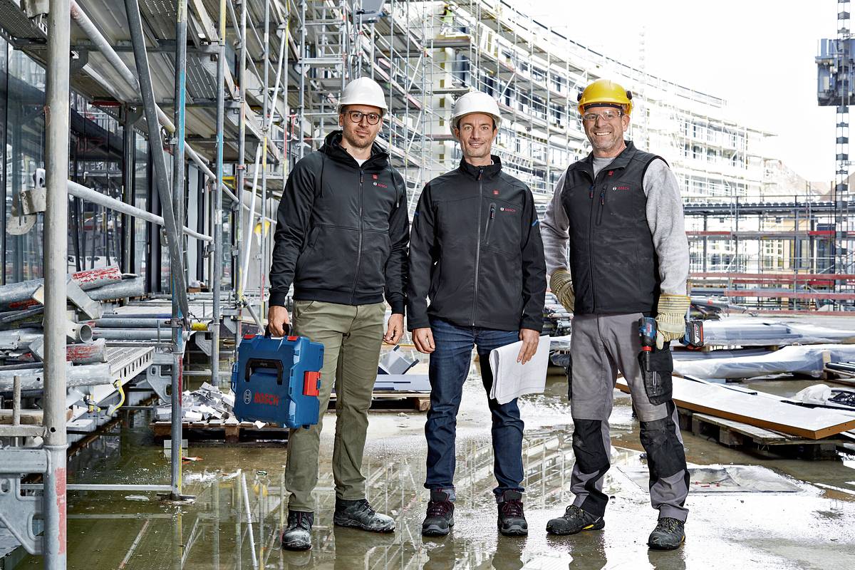 Drei Bauarbeiter in Schutzhelmen stehen auf einer Baustelle vor einem Gebäude im Bau. Sie tragen Arbeitskleidung und halten Werkzeuge.