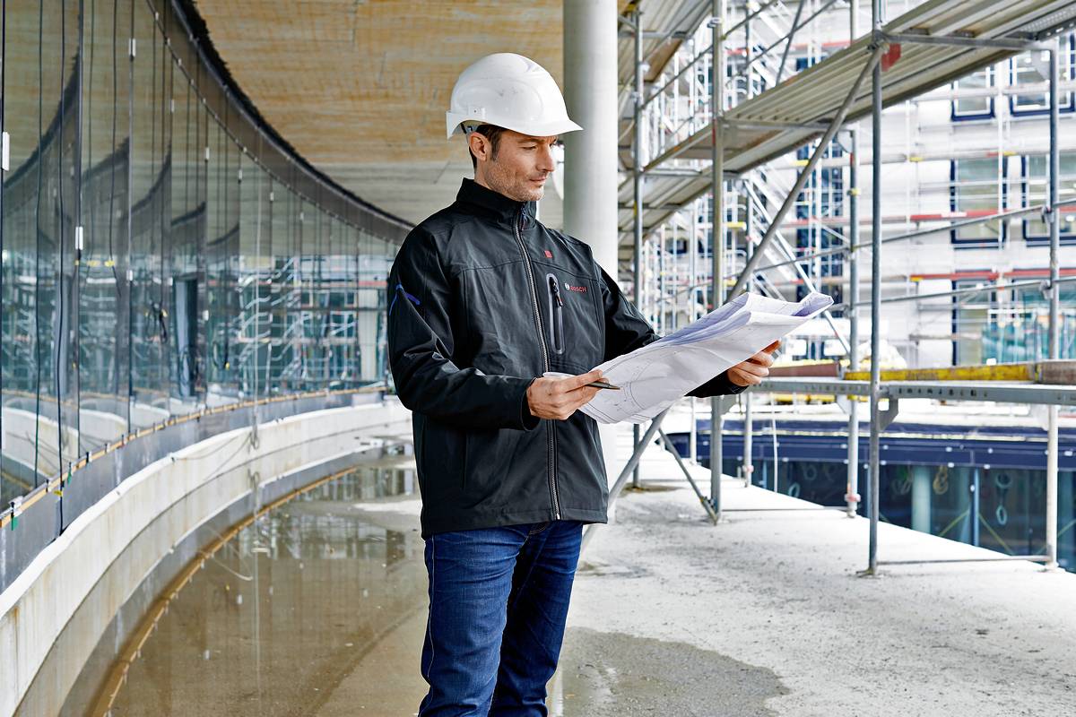 Ein Bauarbeiter mit weißem Helm inspiziert Baupläne auf einer Baustelle. Im Hintergrund ist ein Gerüst und ein modernes Gebäude zu sehen.