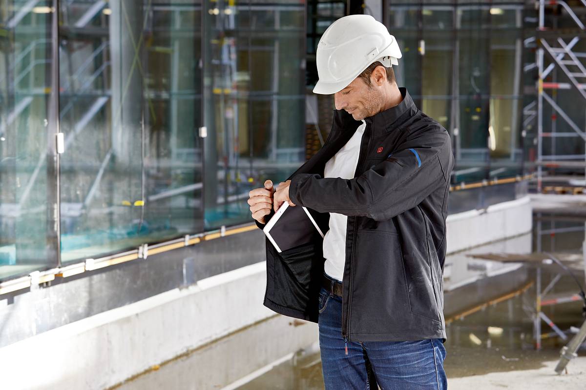 Ein Bauarbeiter mit weißem Helm steht in einem Gebäude im Bau und steckt ein Tablet in seine Jackentasche. Im Hintergrund sind Glasfenster und Baugerüste zu sehen.