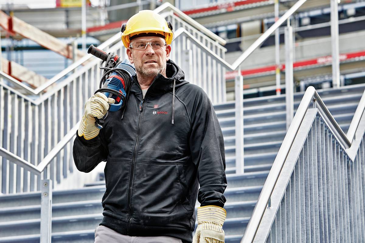 Ein Bauarbeiter mit Schutzhelm und Handschuhen steht auf einer Baustelle und hält ein Elektrowerkzeug. Im Hintergrund sind Baugerüste sichtbar.