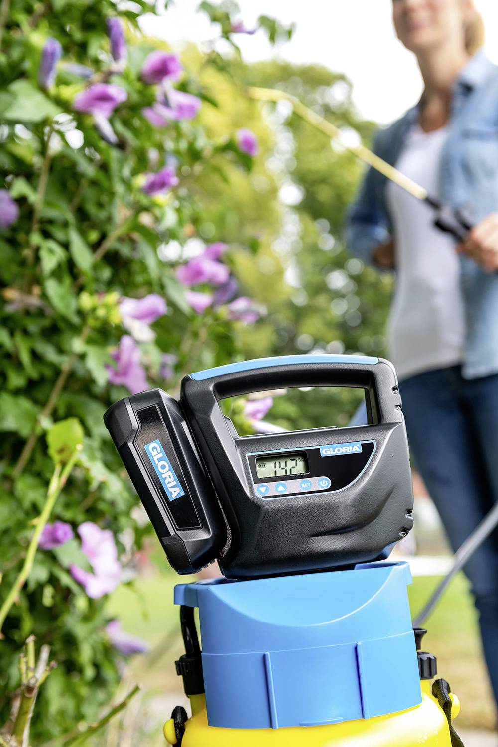 Eine Person sprüht Pflanzen in einem Garten. Im Vordergrund steht eine elektrische Gartenpumpe mit Display für effizientes Arbeiten.