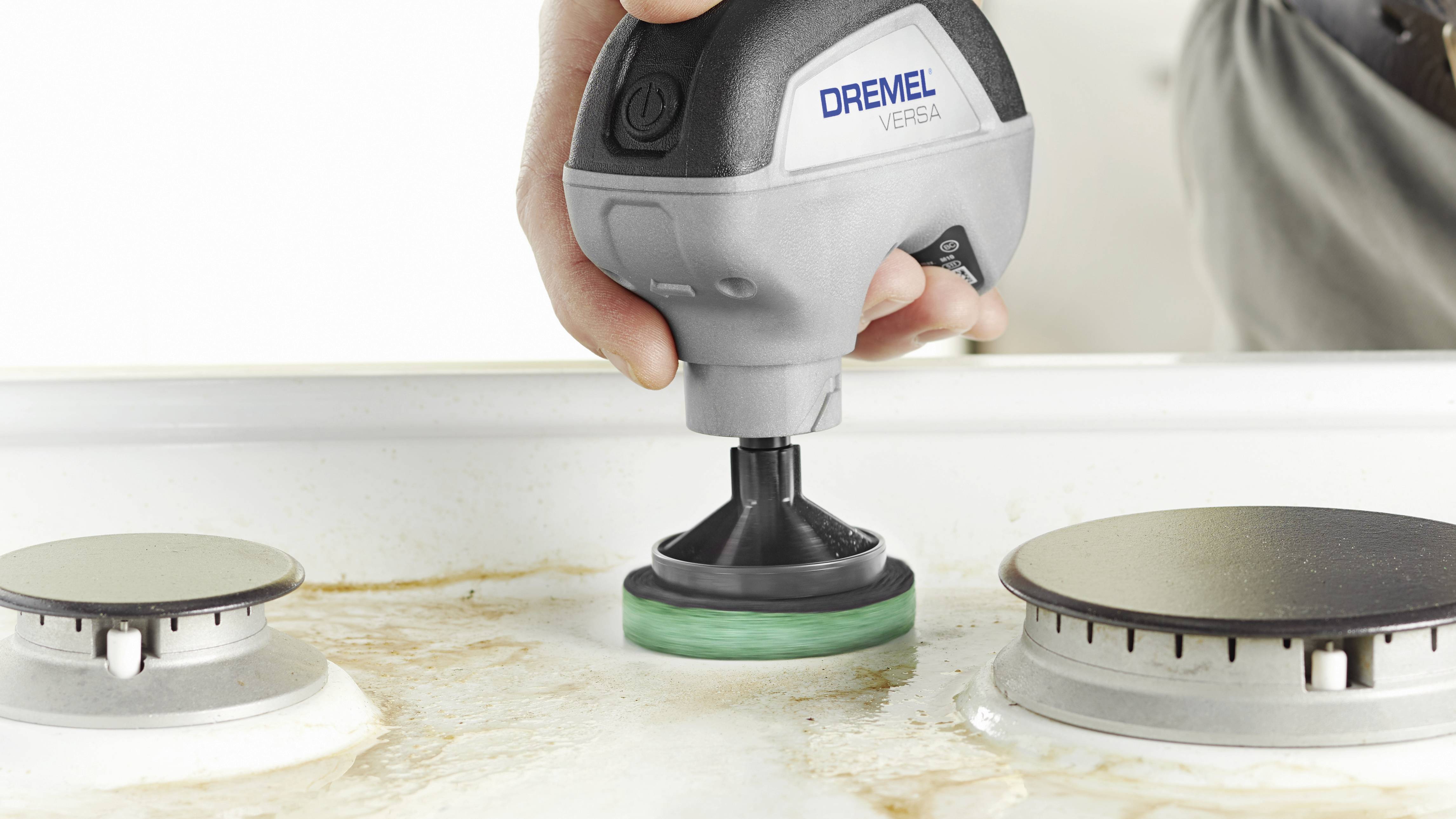 Eine Hand reinigt mit einem elektrischen Reinigungsgerät eine verschmutzte weiße Oberfläche zwischen zwei Herdplatten.