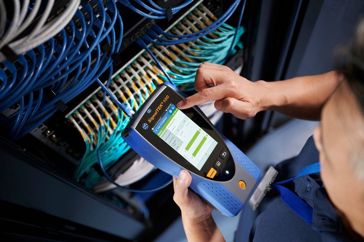 Person bedient ein Netzwerk-Testgerät an einem Server-Rack, um die Netzwerkverbindung zu überprüfen. Kabel sind ordentlich im Rack angeordnet.