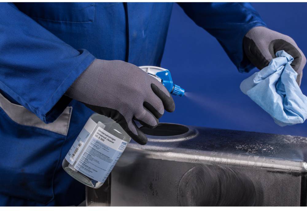 Eine Person in blauer Arbeitskleidung reinigt mit Desinfektionsmittel und Tuch eine metallische Oberfläche.