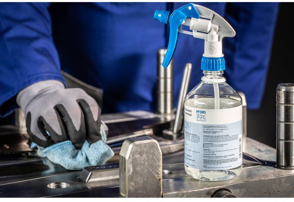 Eine Person mit Handschuhen reinigt eine Metalloberfläche mit einem Tuch. Daneben steht eine Sprühflasche mit Reinigungsmittel.