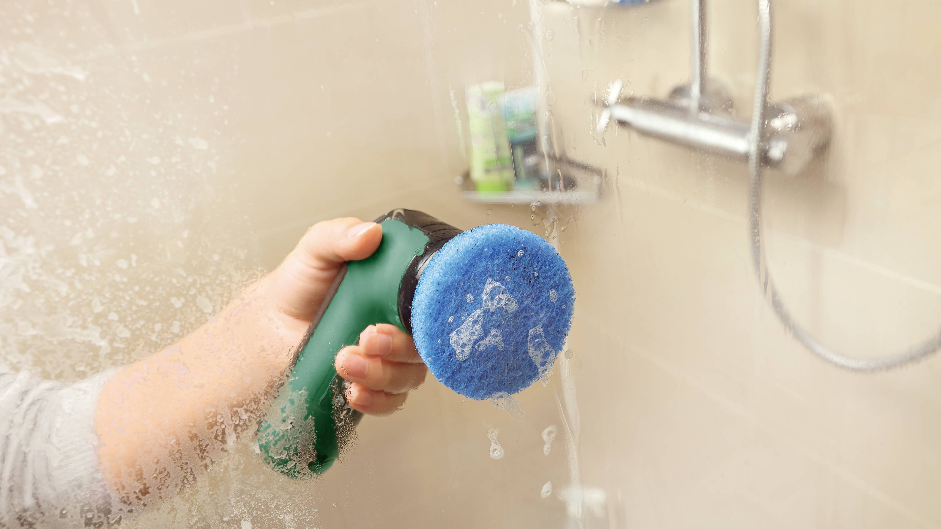 Eine Person reinigt eine Glasscheibe in einer Dusche mit einem blauen Reinigungspad. Wassertropfen sind auf der Scheibe zu sehen.