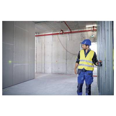 Ein Bauarbeiter in einer Sicherheitsweste und einem Helm verwendet ein Lasernivellement, um in einem teilweise fertiggestellten Betonraum mit freigelegten Rohrleitungen zu vermessen.