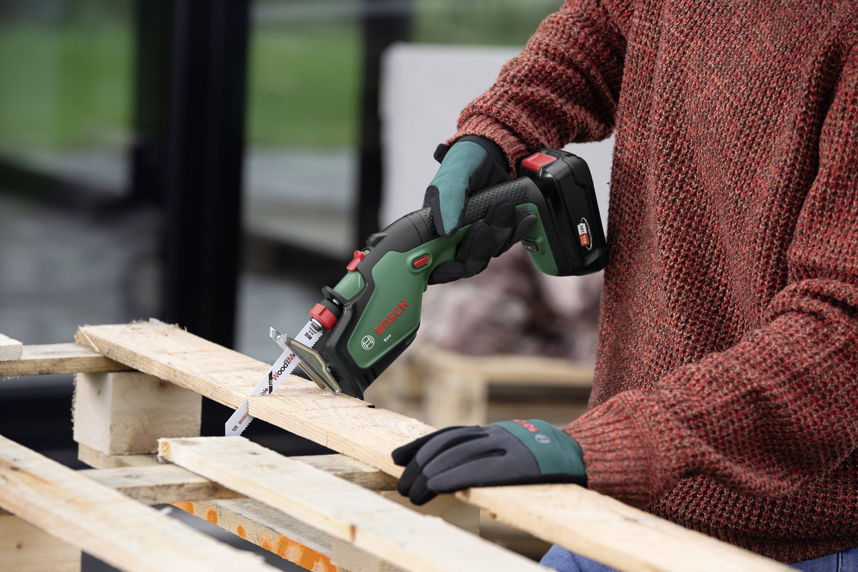 Eine Person schneidet Holz mit einer elektrischen Stichsäge. Sie trägt einen roten Pullover und Arbeitshandschuhe.