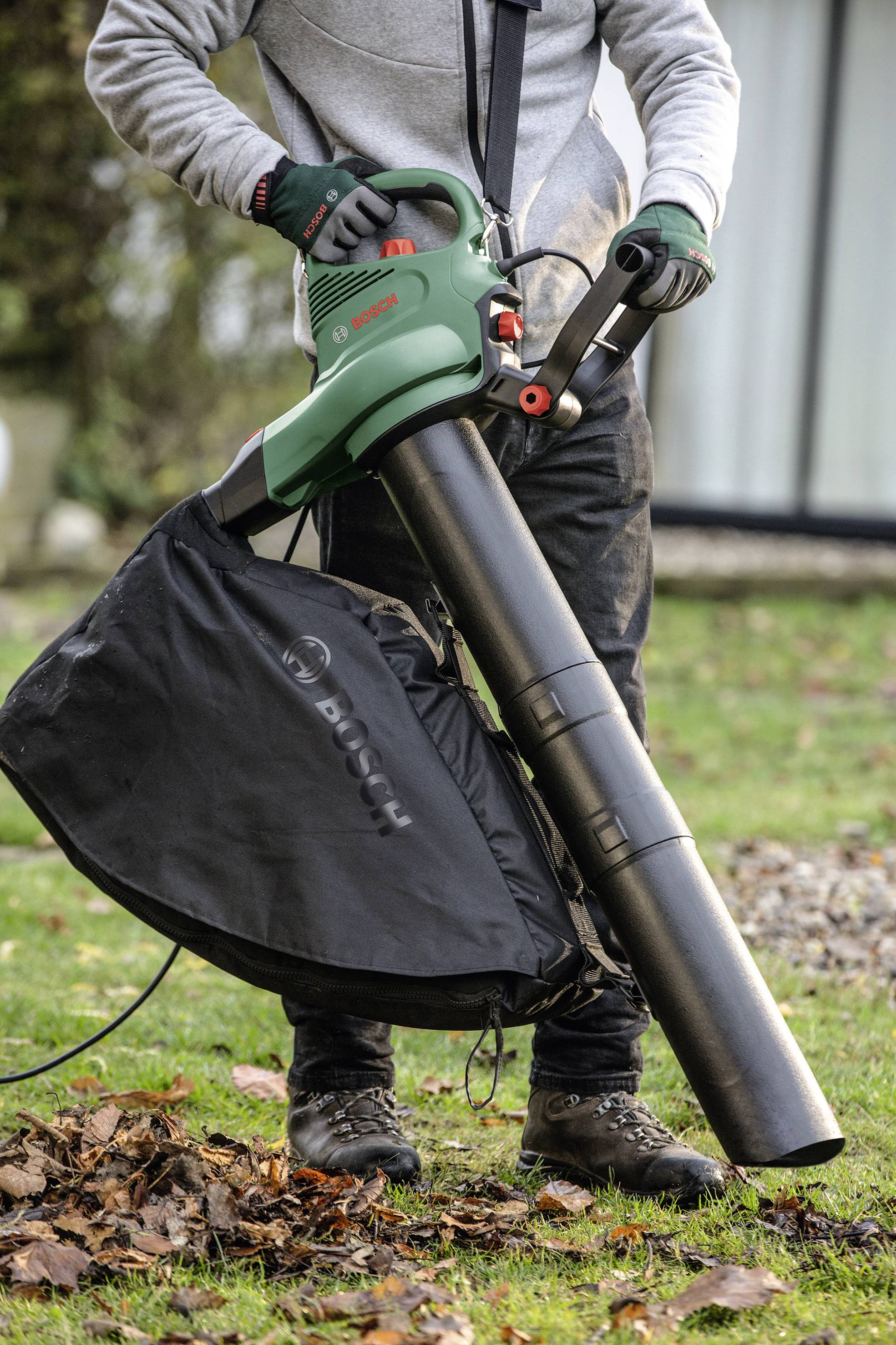 Person benutzt einen Laubbläser, um Blätter auf einem Rasen zu entfernen. Der Laubbläser hat einen Fangbeutel und ist grün-schwarz.