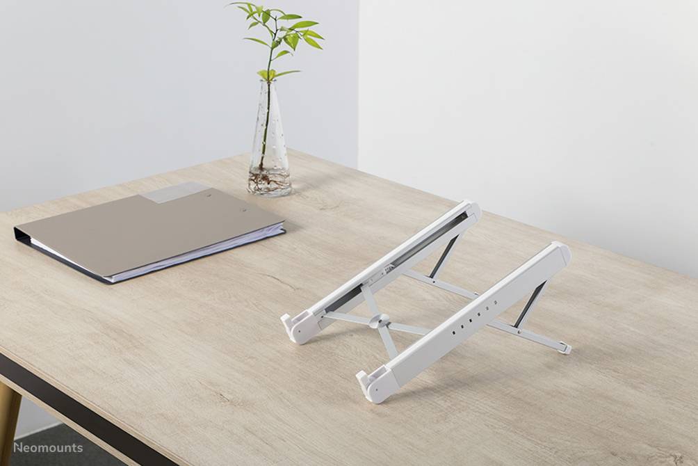 Ein Laptop-Ständer aus Metall steht auf einem Holztisch neben einer Pflanze in einer Glasvase und einem geschlossenen Notizbuch.