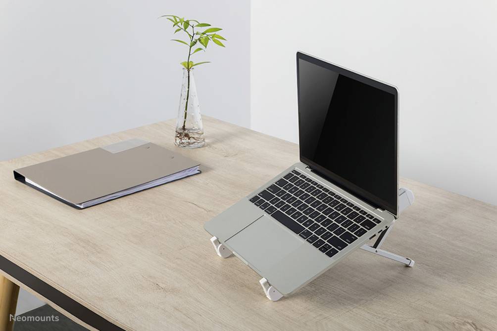 Schreibtisch mit Laptop auf einem höhenverstellbaren Ständer, daneben eine kleine Pflanze in einer Vase und ein geschlossener Ordner.