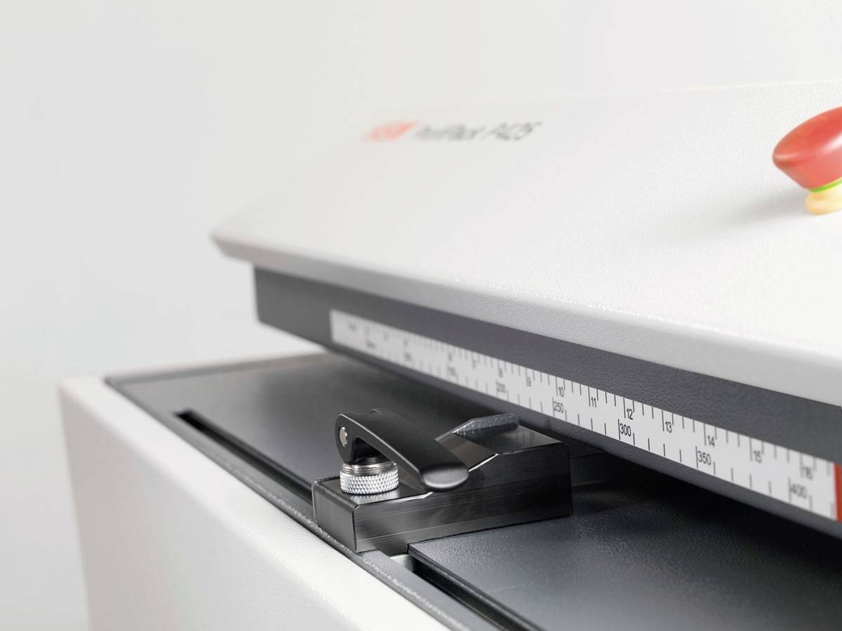 'Papier-Schneidemaschine mit Messskala und rotem Sicherheitsknopf auf der Oberseite. Eignet sich für präzise Papierschnitte in Büro oder Druckerei.'