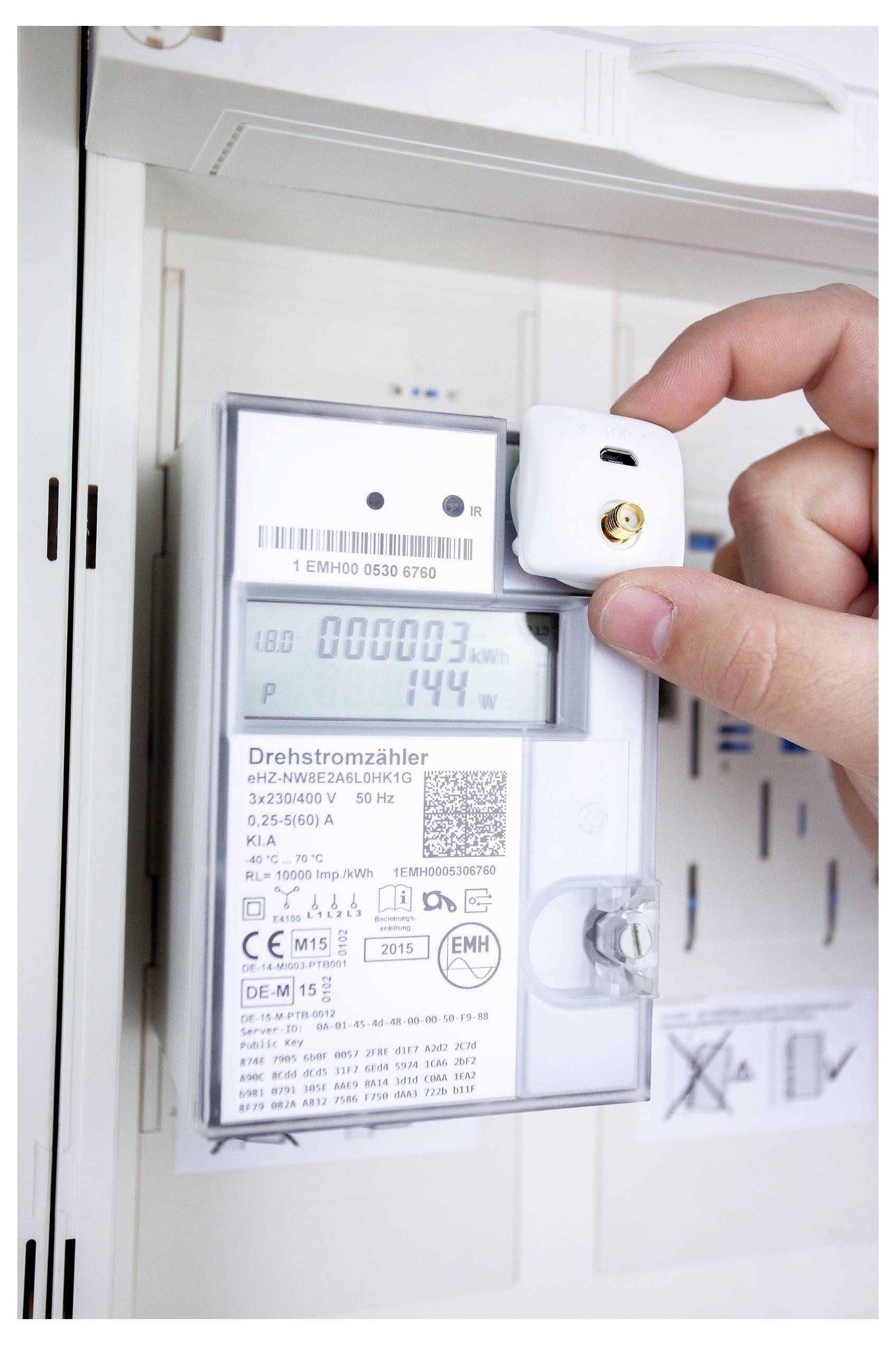Eine Hand, die einen kleinen Sensor auf einem Stromzähler platziert, der '000003' kWh und '144' Watt anzeigt und damit die Energieverbrauchsüberwachung dokumentiert.