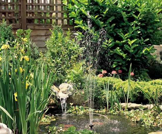 Ein kleiner Garten mit einem Teich, sprudelnder Fontäne und üppiger Vegetation. Im Hintergrund sind Holzwände und Blumen zu sehen.