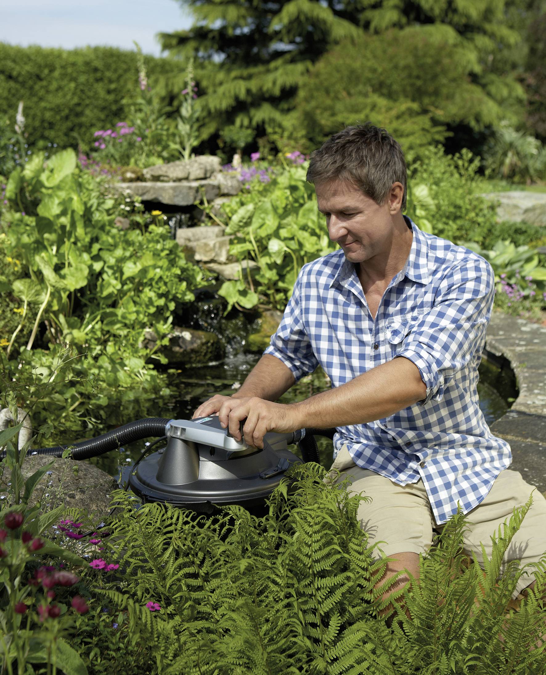Ein Mann in kariertem Hemd sitzt im Garten vor einem Teich und bedient ein Gerät. Im Hintergrund sind grüne Pflanzen und ein Wasserfall.