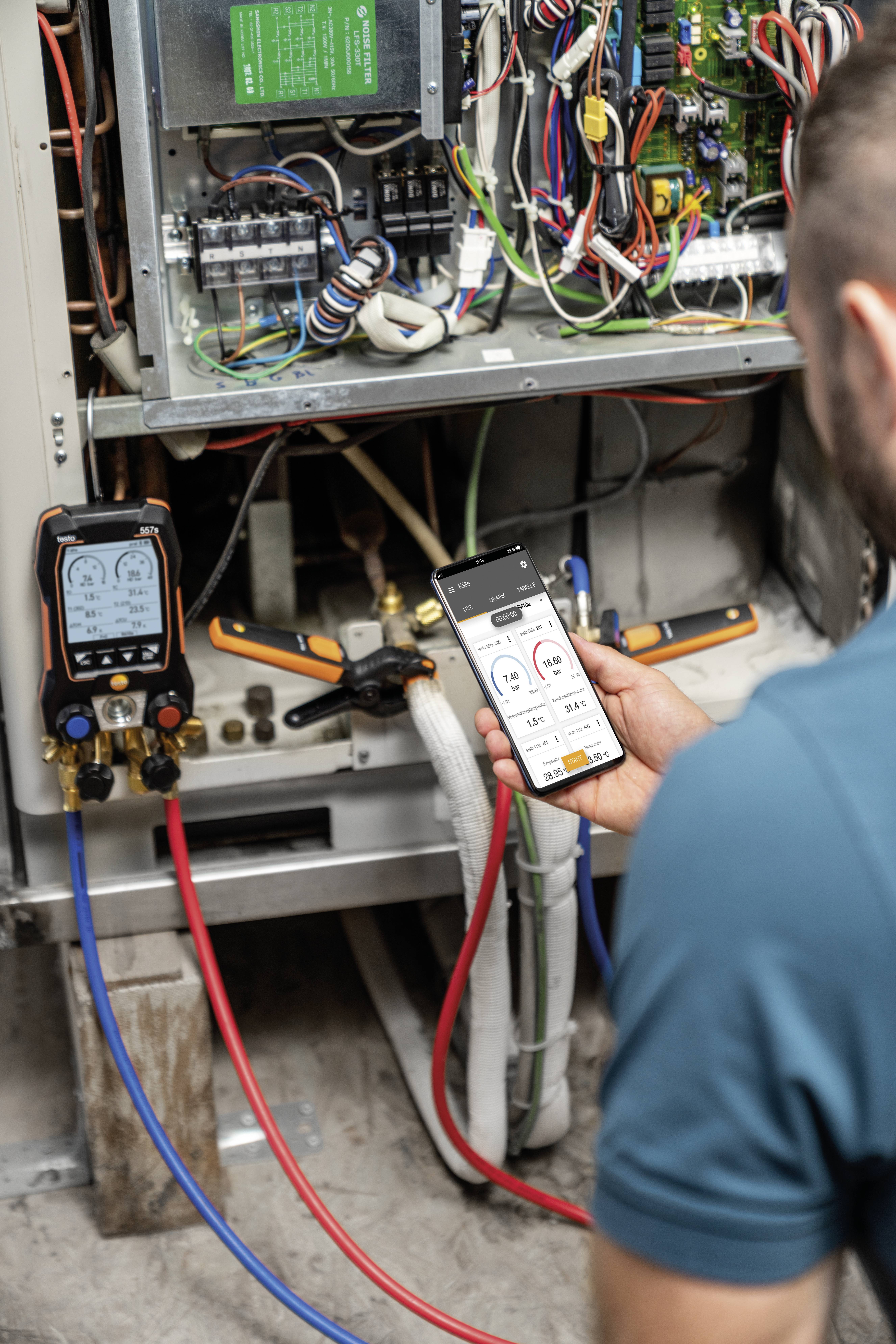 Eine Person überprüft ein Klimagerät mit einem Smartphone und einem Diagnosegerät. Kabel und Schaltungen sind sichtbar.