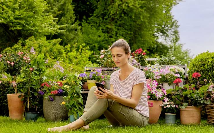 Eine Person sitzt auf Gras mit einem Smartphone, umgeben von verschiedenen Topfblumen und üppiger Vegetation, was eine friedliche Gartenkulisse schafft.