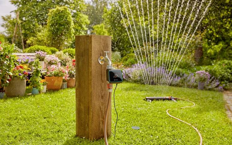 Eine Gartenszene, die ein intelligentes Bewässerungssystem mit einem digitalen Timer zeigt, der an einen Schlauch angeschlossen ist und üppige Blumen und Grünpflanzen bewässert.