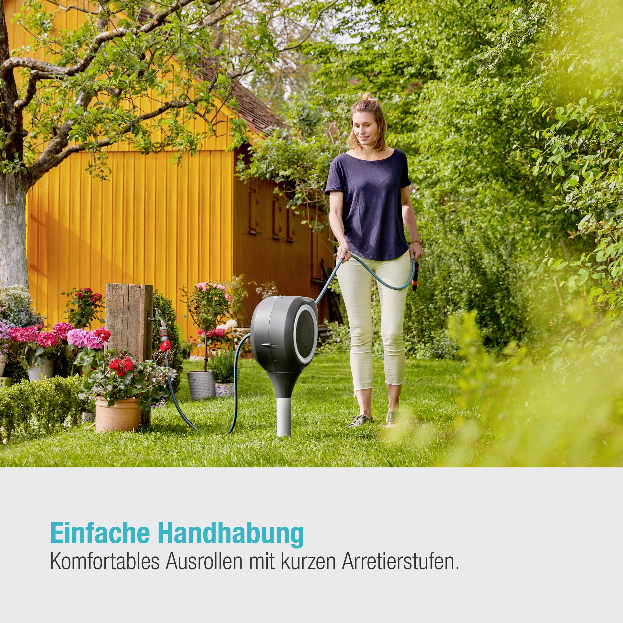 Eine Frau steht im Garten neben einem gelben Haus und benutzt eine Schlauchtrommel, um Pflanzen zu bewässern. Text: 'Einfache Handhabung'.