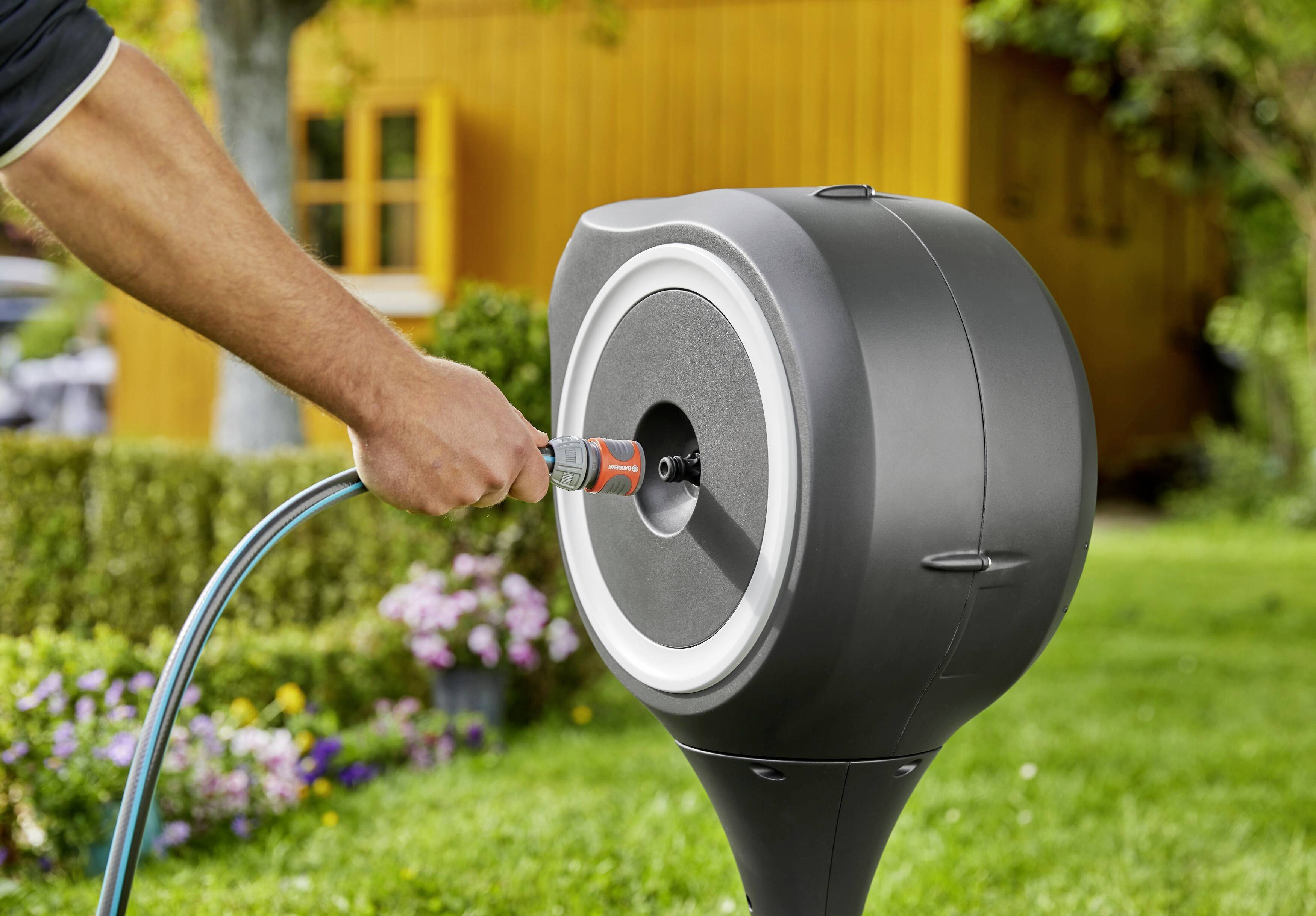 Eine Person verbindet einen Gartenschlauch mit einer schwarzen Schlauchtrommel im Garten. Im Hintergrund ist ein gelbes Gebäude.