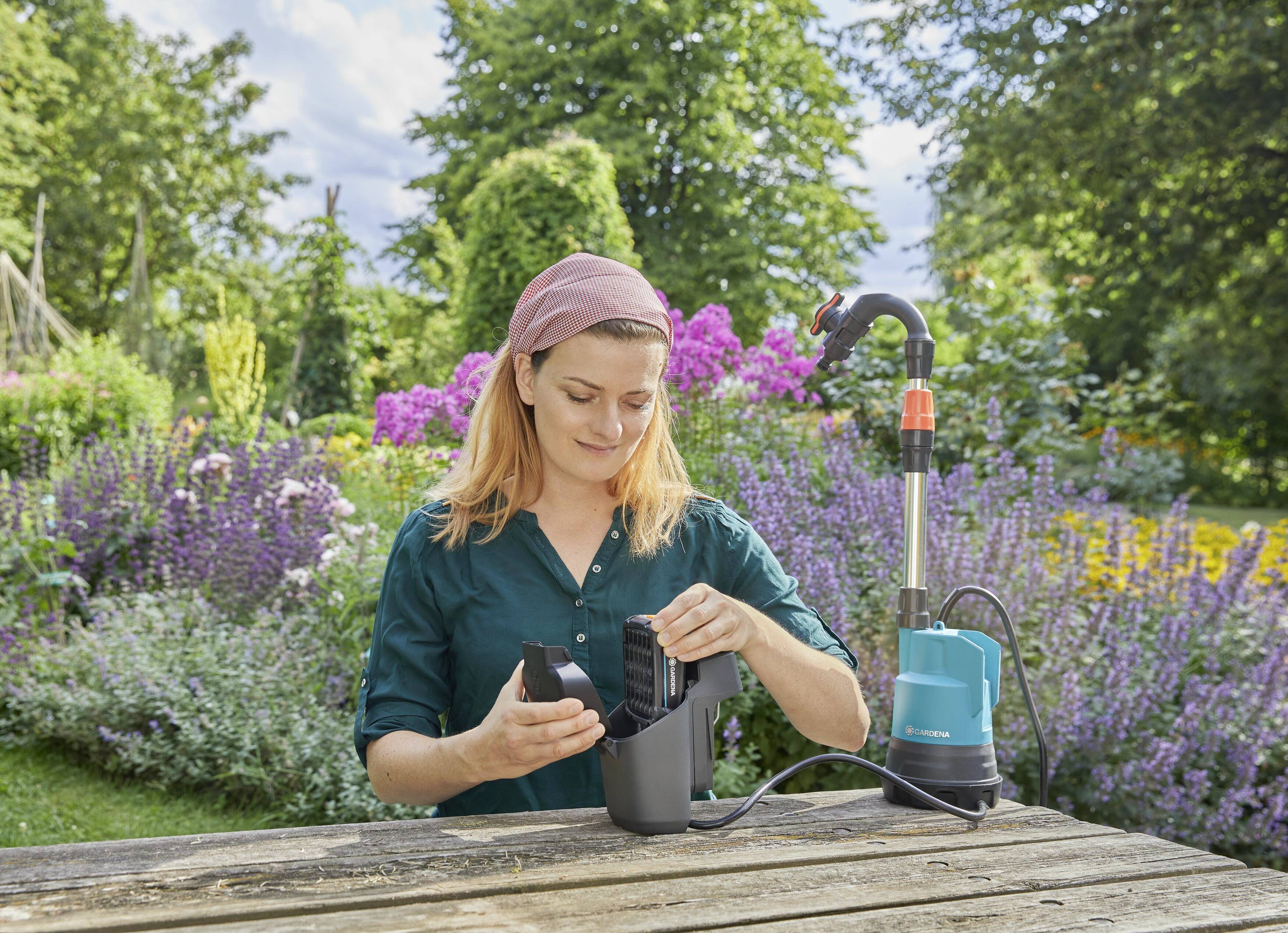 Frau im Garten verwendet eine blaue Wasserpumpe auf einem Holztisch, umgeben von bunten Blumen und Bäumen.