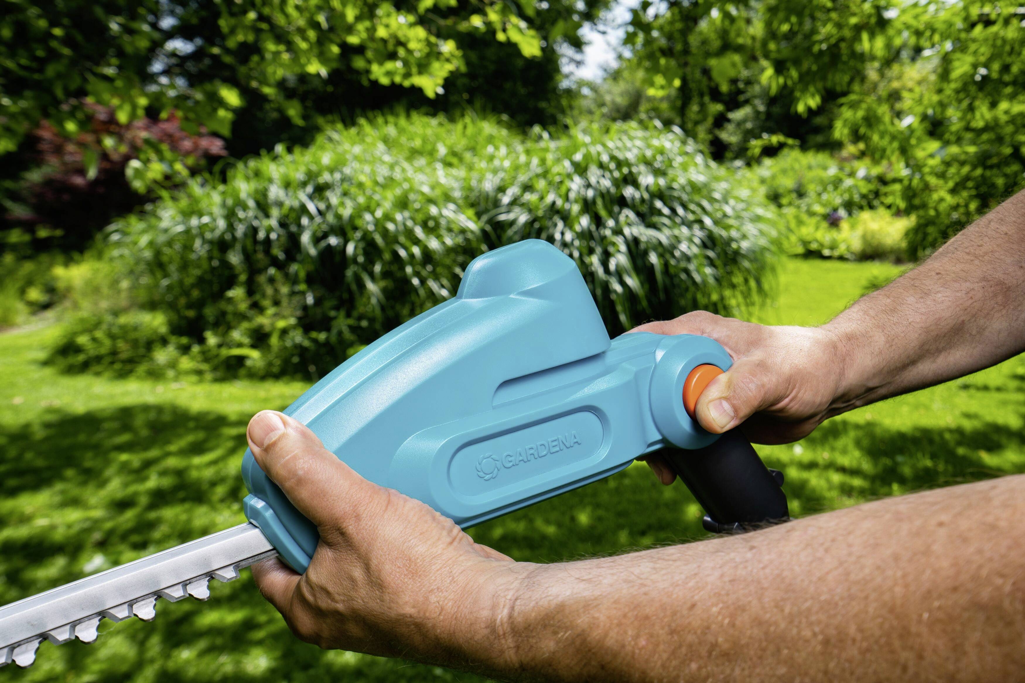 Person hält eine elektrische Heckenschere im sonnigen Garten. Die Schere ist blau und wird mit beiden Händen bedient. Im Hintergrund sind grüne Büsche.