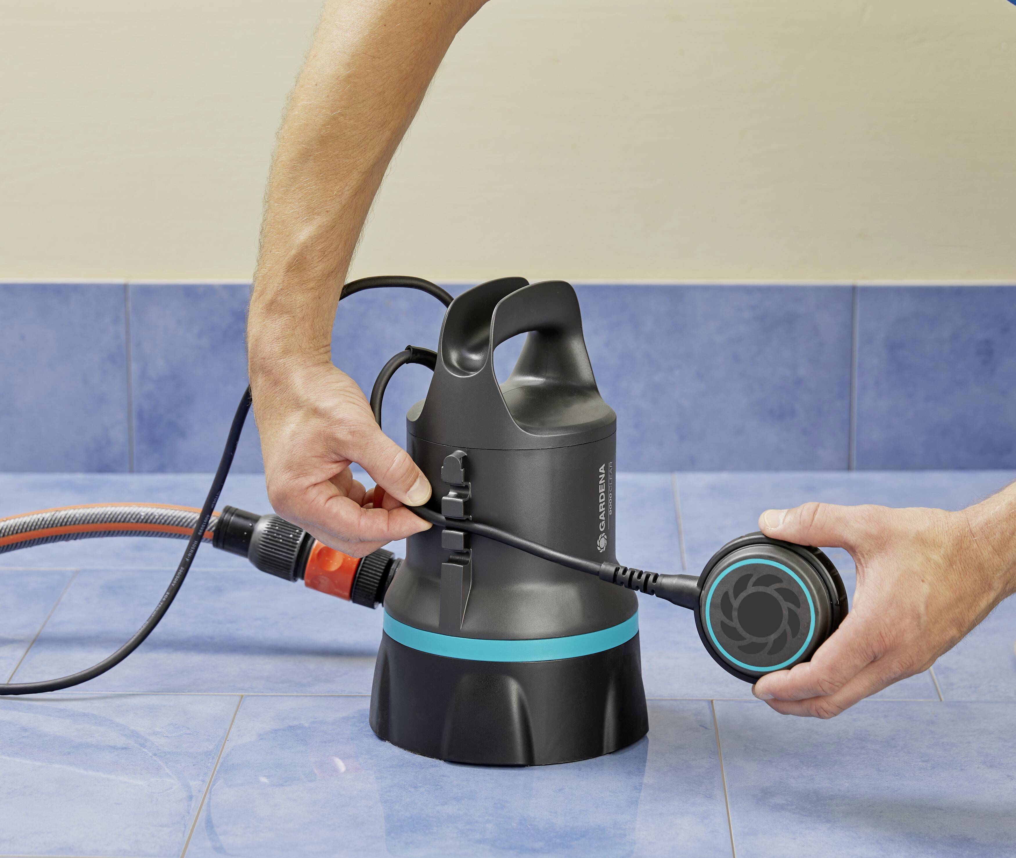 Eine Person hält und prüft eine schwarze Tauchpumpe mit einem blauen Streifen, die auf blauen Fliesen steht.