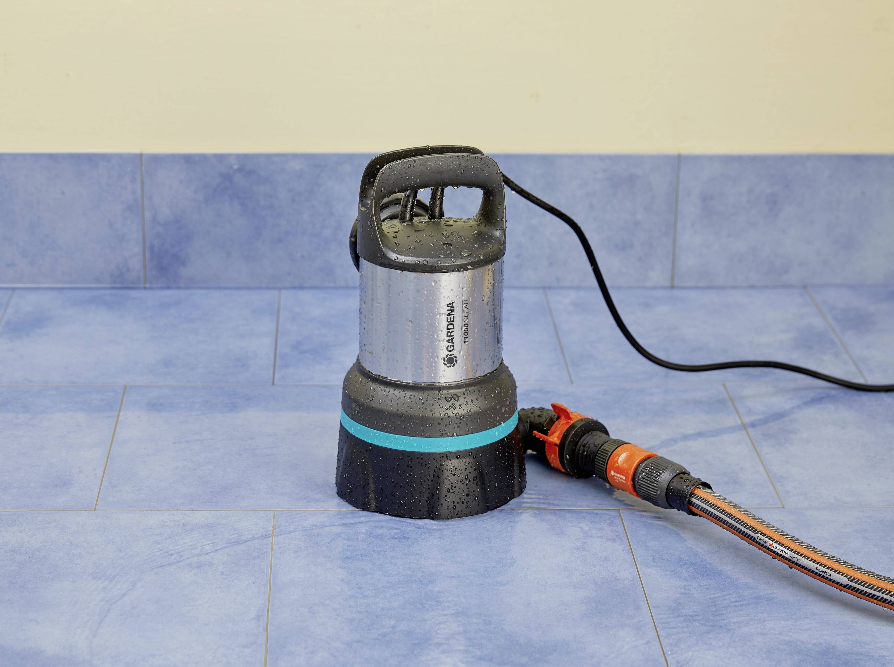 Eine silberne Tauchpumpe steht auf einem gefliesten Boden. Ein schwarzes Stromkabel mit orangener Steckverbindung führt von der Pumpe weg.