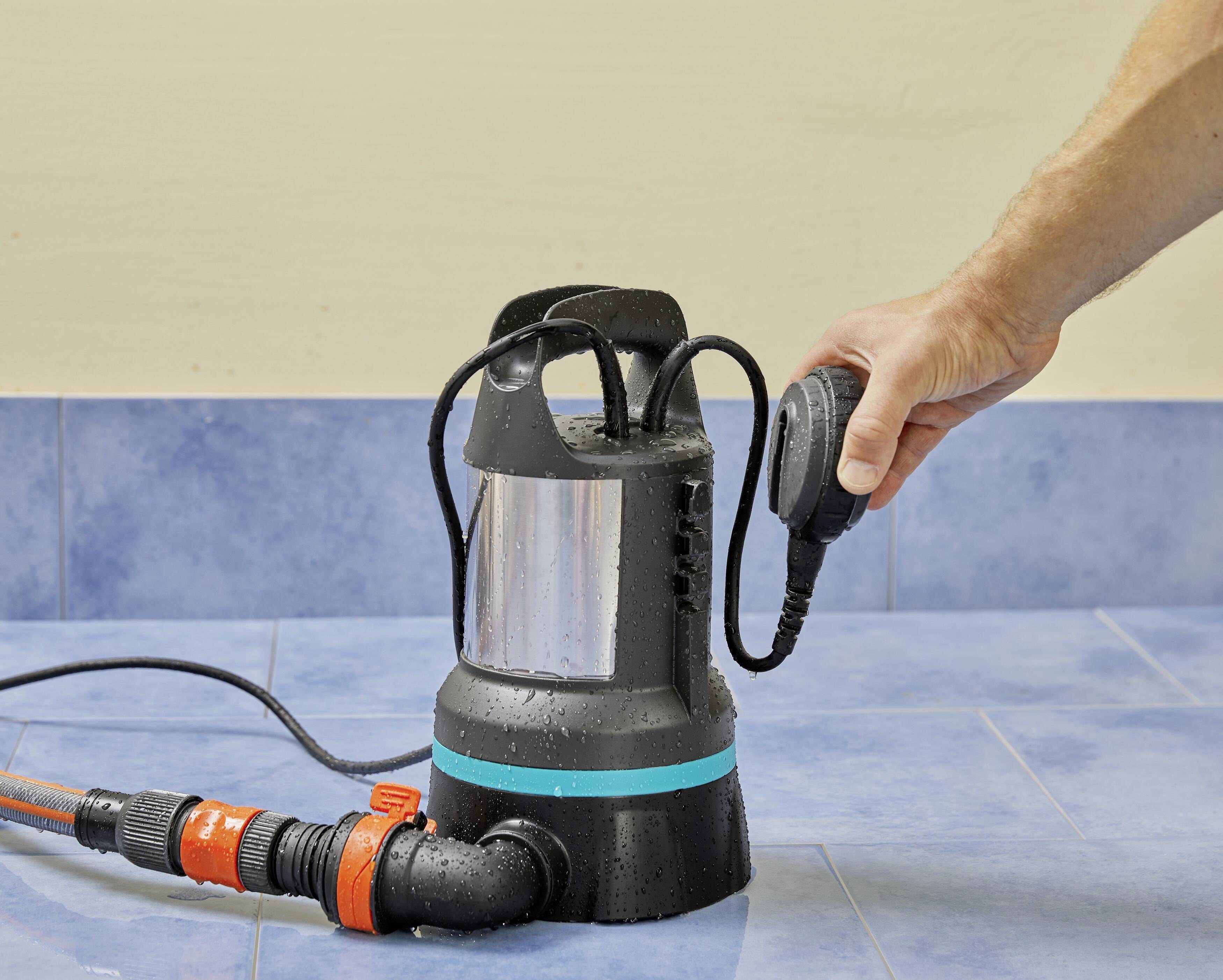 Eine Hand hält einen Schwimmschalter, der mit einer Tauchpumpe verbunden ist. Die Pumpe steht auf gefliestem Boden.