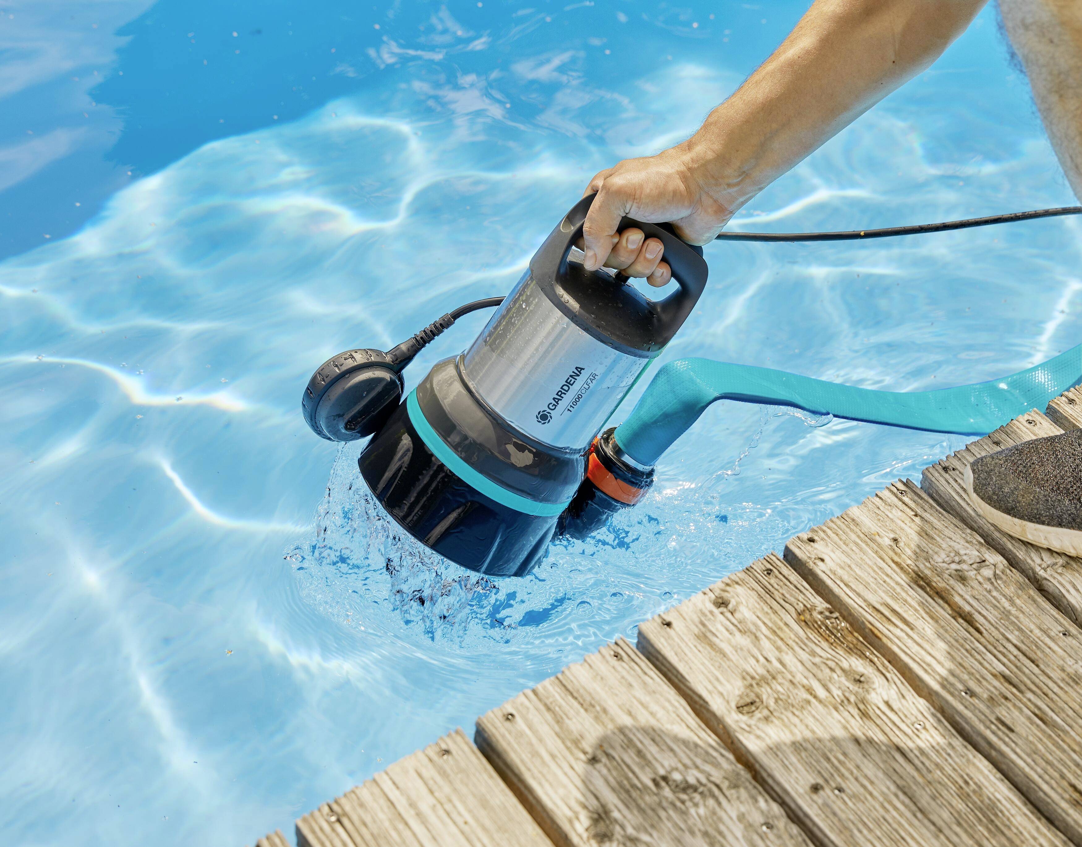 Eine Person hält eine Tauchpumpe, die Wasser aus einem Pool pumpt. Das blaue Wasser und ein Holzdeck sind sichtbar.