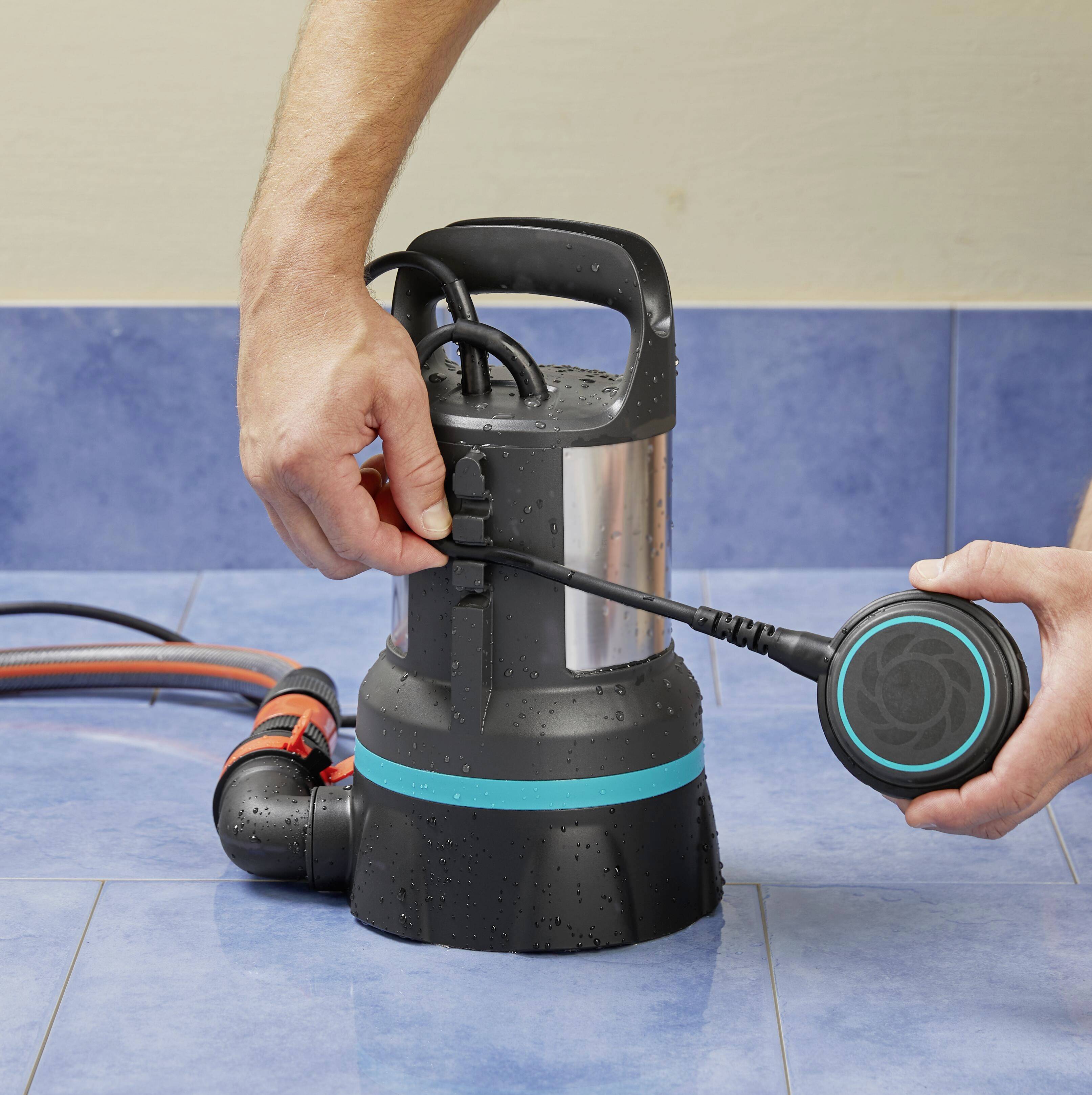 Eine Person hält eine schwarze Wasserpumpe mit blauem Streifen auf blauem Fliesenboden und steckt ein Kabel ein.