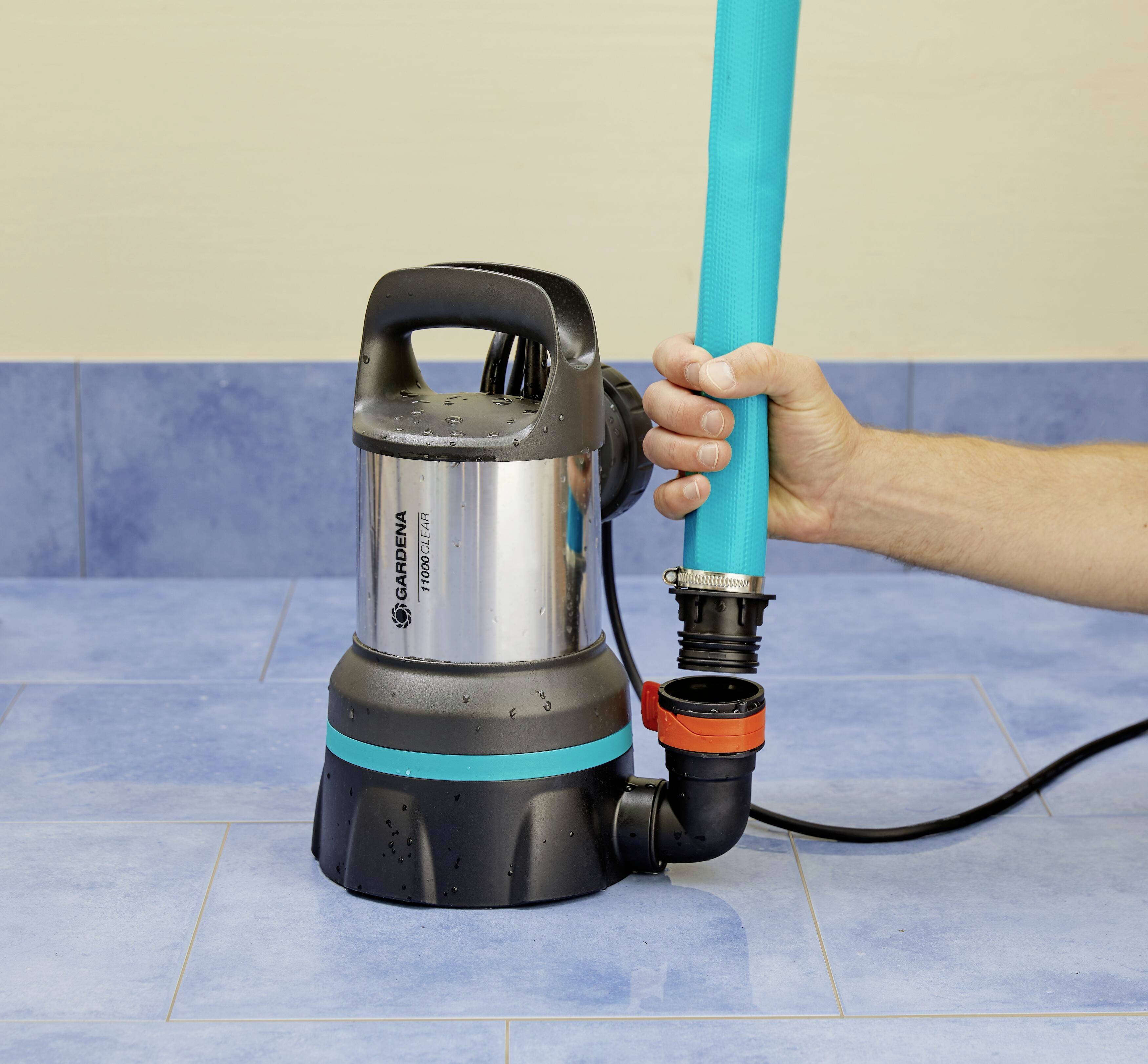 Hand hält Wasserpumpe mit blauem Schlauch auf blau gefliestem Boden. Gardena Pumpe bereit zum Abpumpen von Wasser.